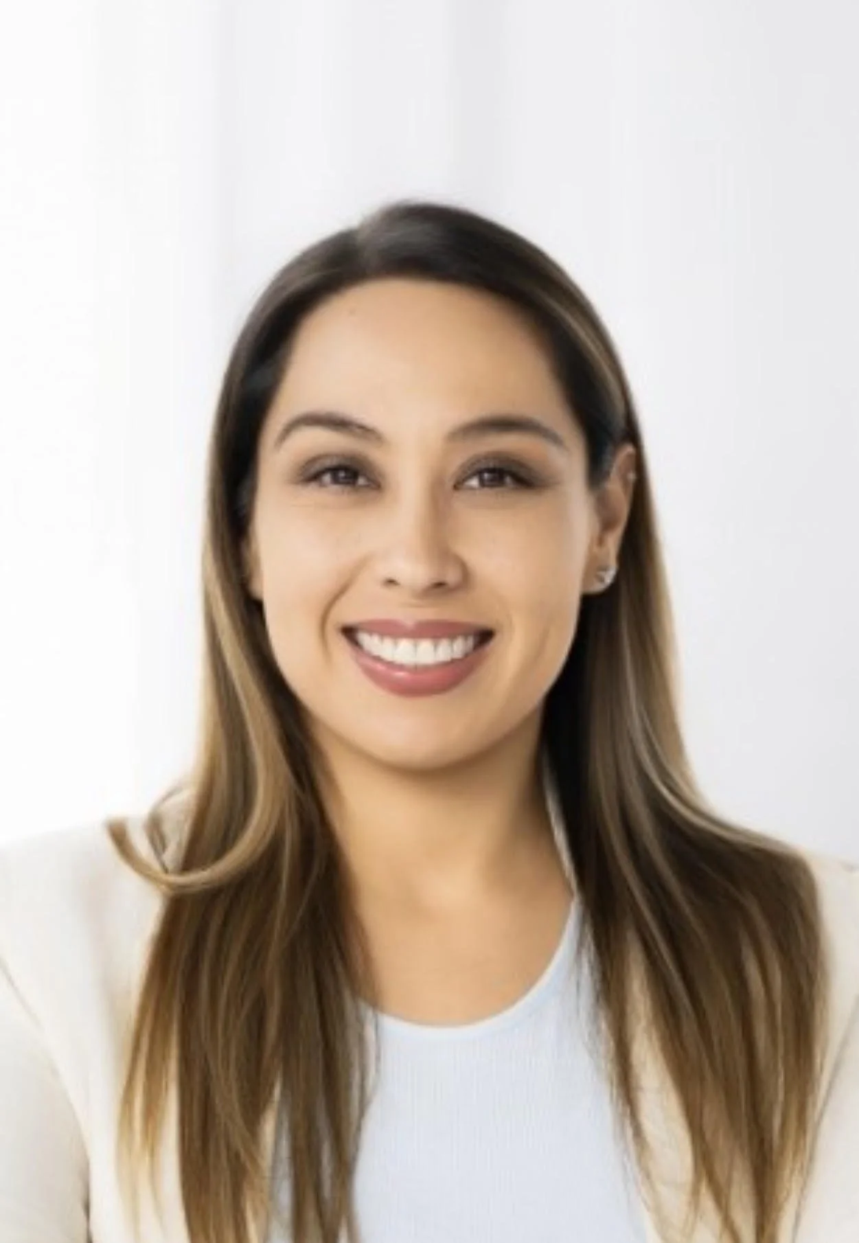 A young woman with long, wavy brown hair smiling at the camera, wearing a white top and beige blazer, standing in front of a white background.