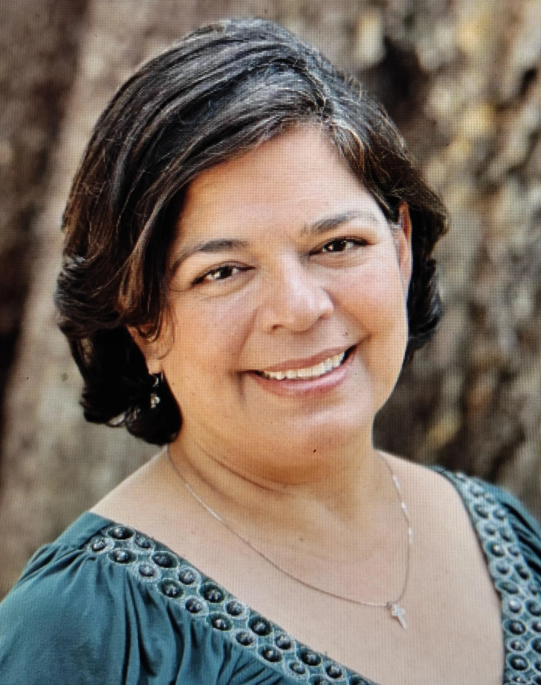 Close-up of a woman with short dark hair smiling, wearing a blue dress with embroidered details, a silver necklace with a cross pendant, and a delicate chain necklace.