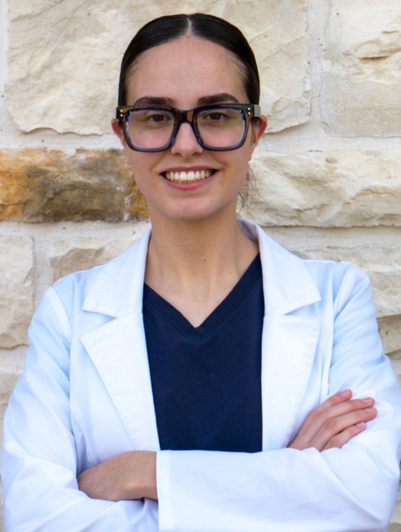 A woman with dark hair, wearing large glasses, a white lab coat, and a dark shirt, standing in front of a beige stone wall, smiling with her arms crossed.
