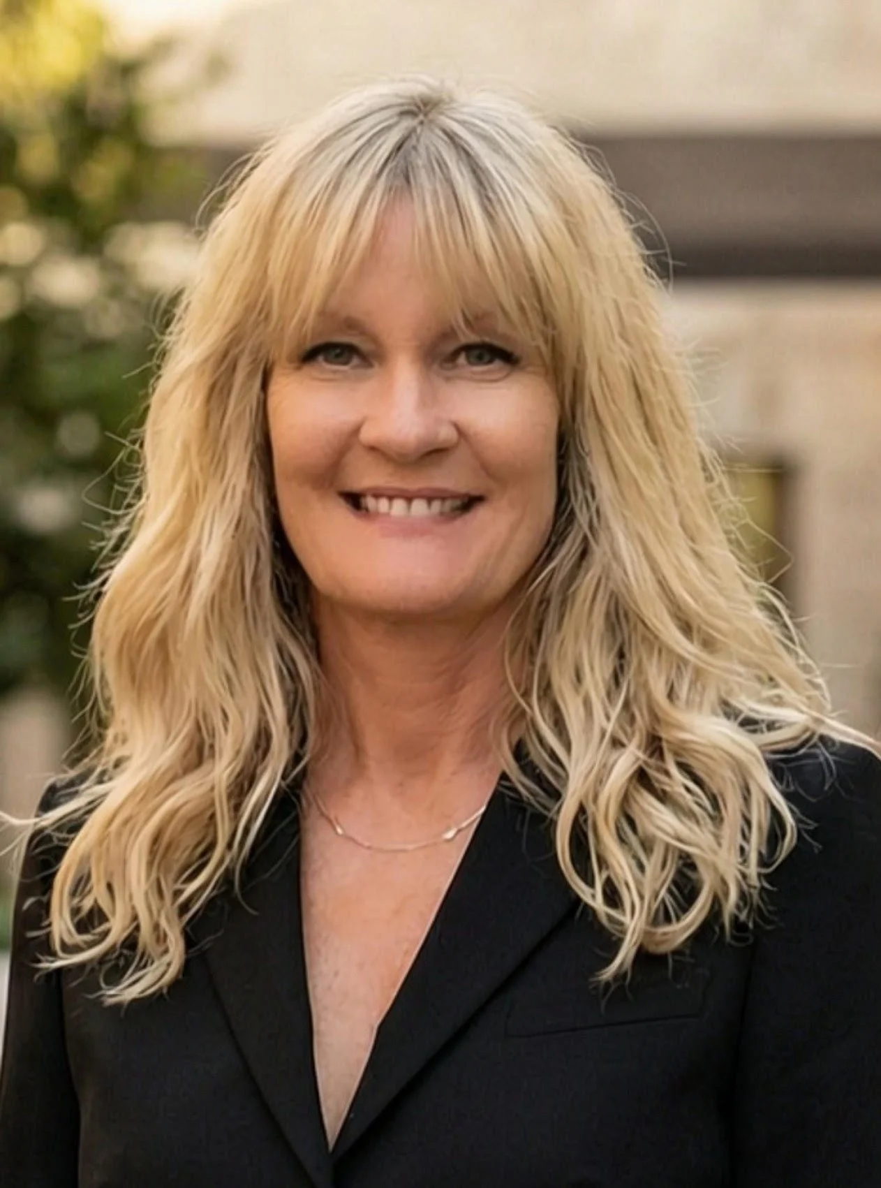 A woman with long, wavy blonde hair, smiling, wearing a black blazer and a delicate necklace.