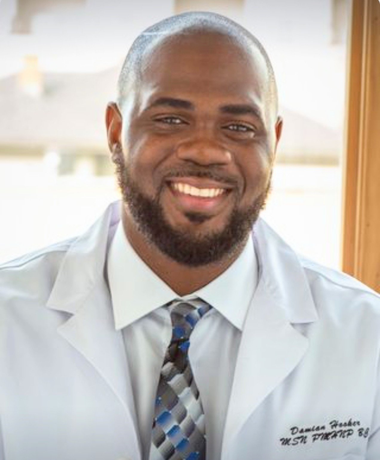 Portrait of a smiling man wearing a white medical coat and a patterned tie, sitting indoors near a window.