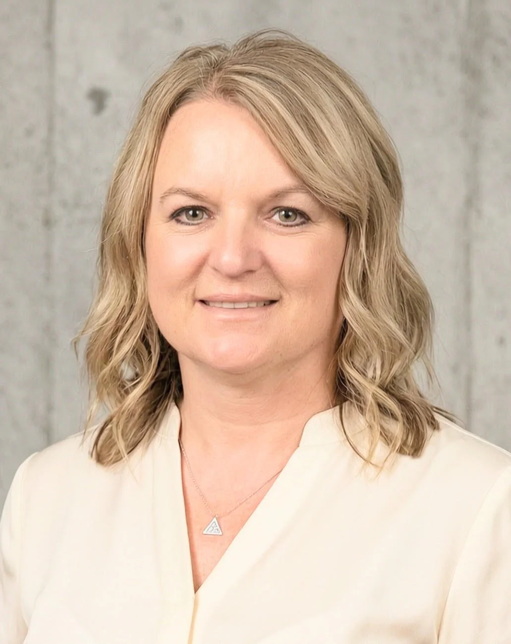 Professional woman with shoulder-length blonde hair, wearing a white top and a triangular necklace, smiling in front of a textured gray wall.