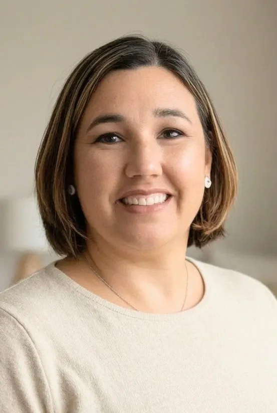 A woman with short brown hair smiling, wearing a light-colored top and earrings, sitting in a room with neutral-colored walls and a lamp in the background.
