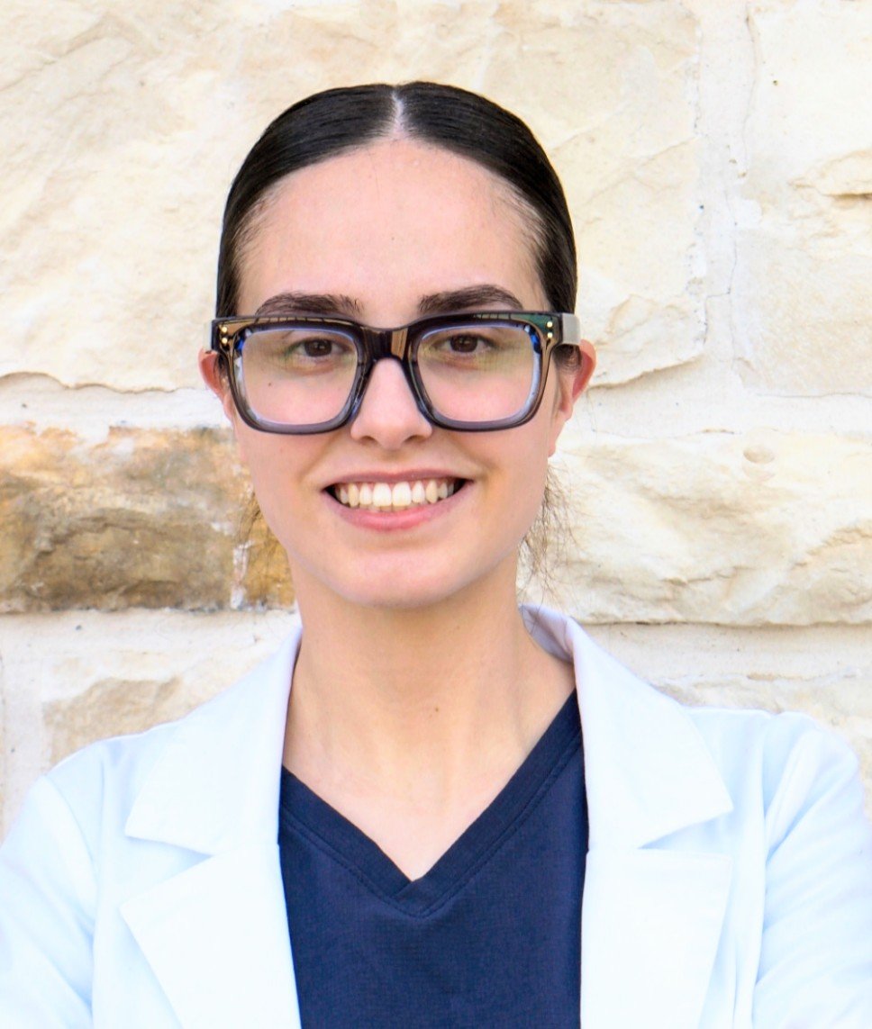 A woman with dark hair and large glasses smiling in front of a stone wall.