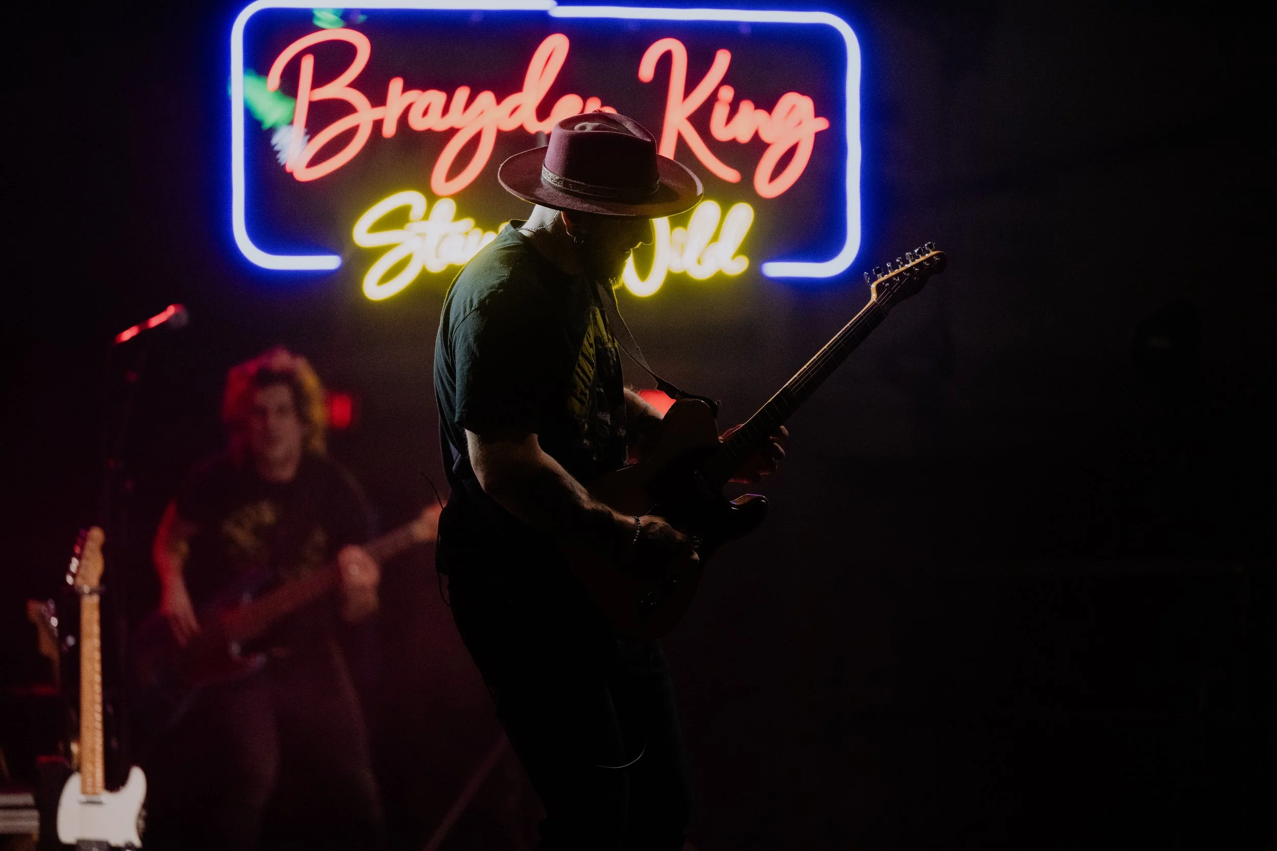 A musician wearing a hat playing an electric guitar on stage, silhouetted against a neon sign that reads 'Brayden King Streaming Wild' in a dark setting.