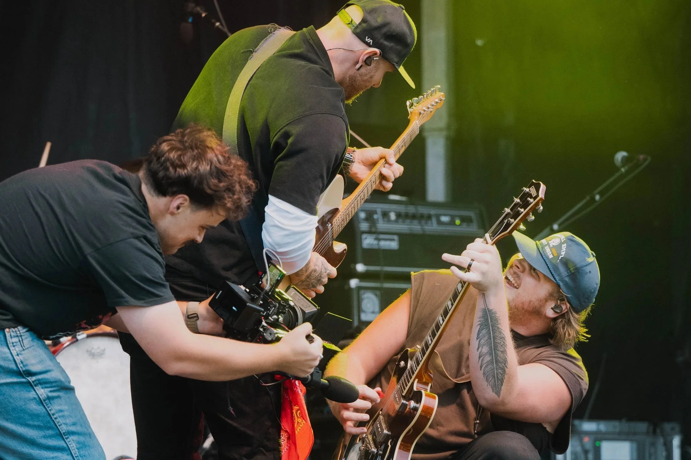 Three musicians on stage, two playing guitars and one holding a camera, with stage equipment in the background.