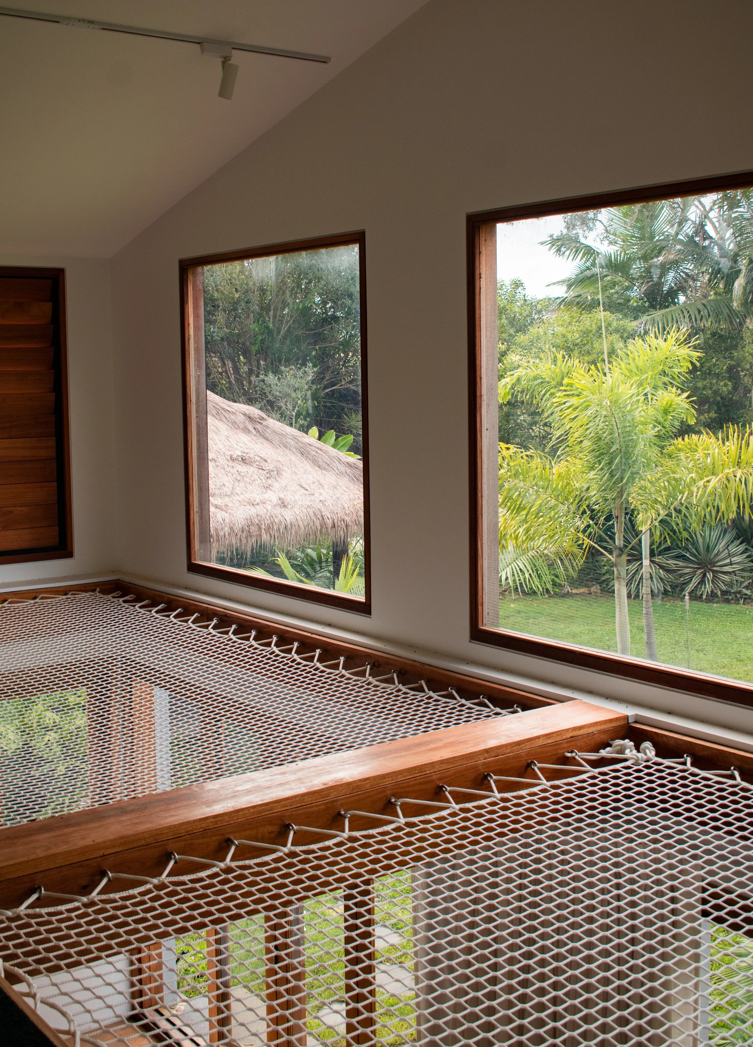 Interior of a room with large windows revealing lush green tropical plants outside. A hammock made of netting and wooden frame is in the foreground, with a thatched roof structure visible outside through the windows.