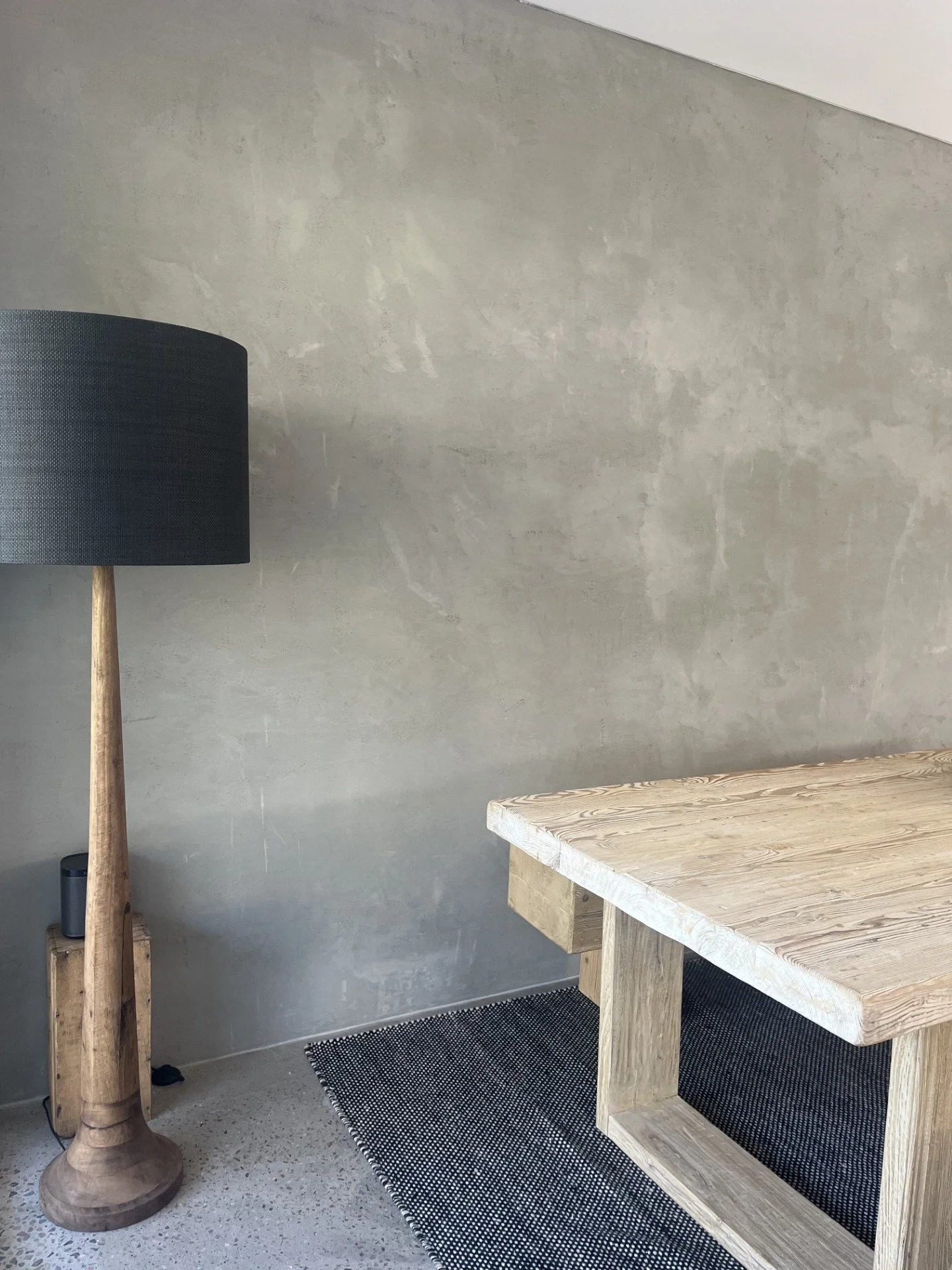 A corner of a room with a wooden table, a black and gray rug, and a tall black lamp with a wooden base.