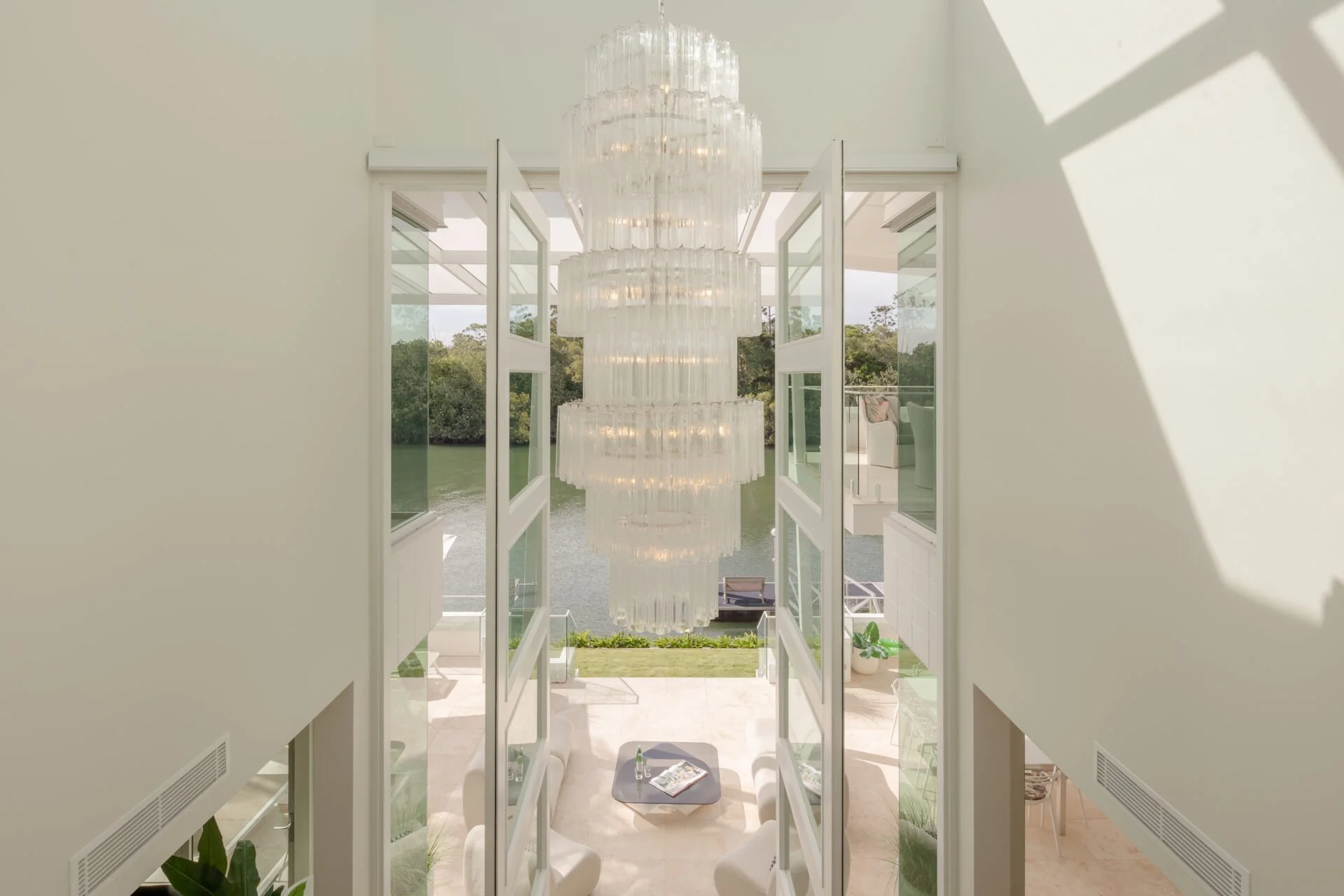 A view through open glass doors into a bright room with a large crystal chandelier hanging from the ceiling, overlooking a lake and greenery outside.