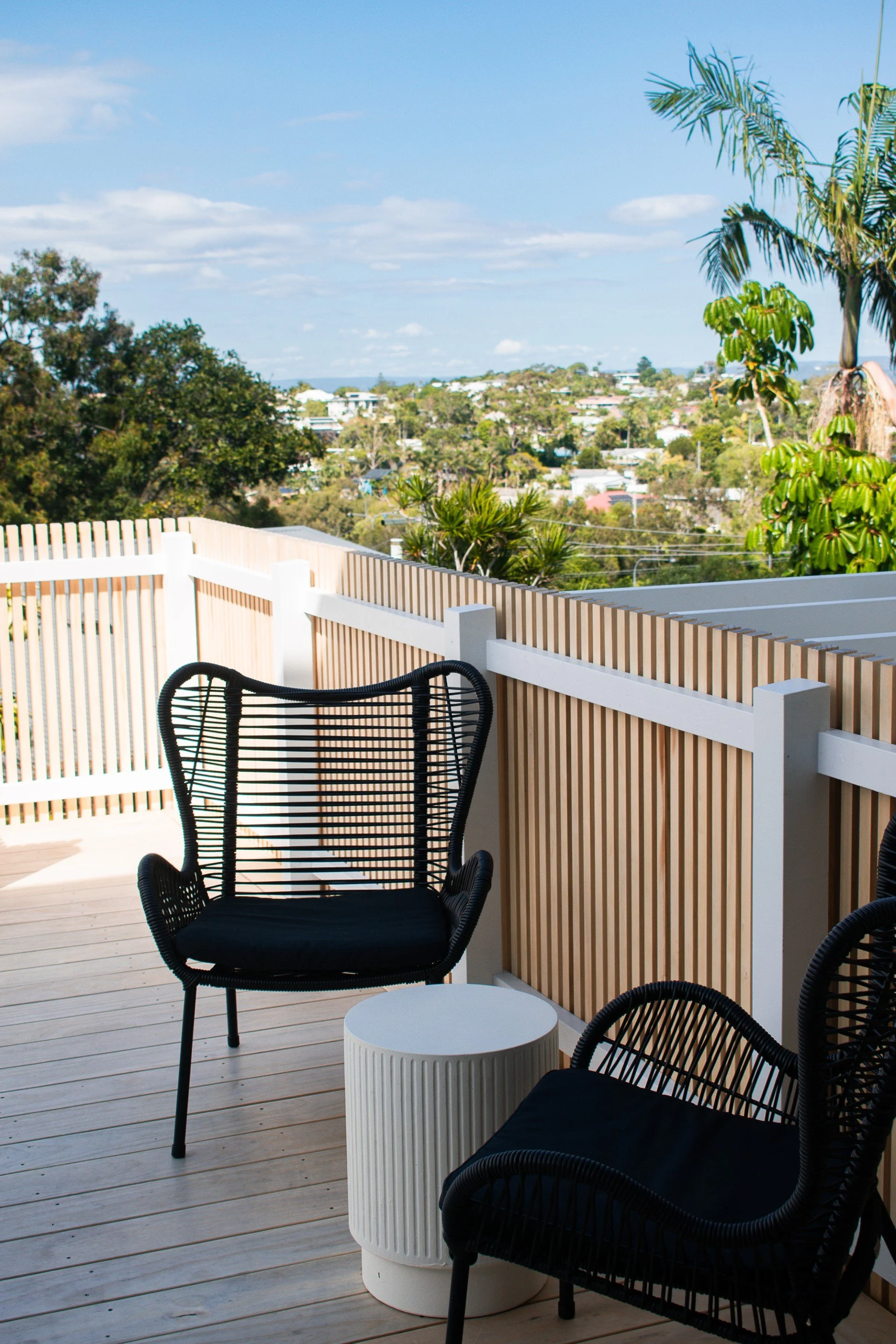 An outdoor balcony with black chairs, a white round side table, a wooden fence, and a view of trees and houses in the distance under a blue sky with scattered clouds.