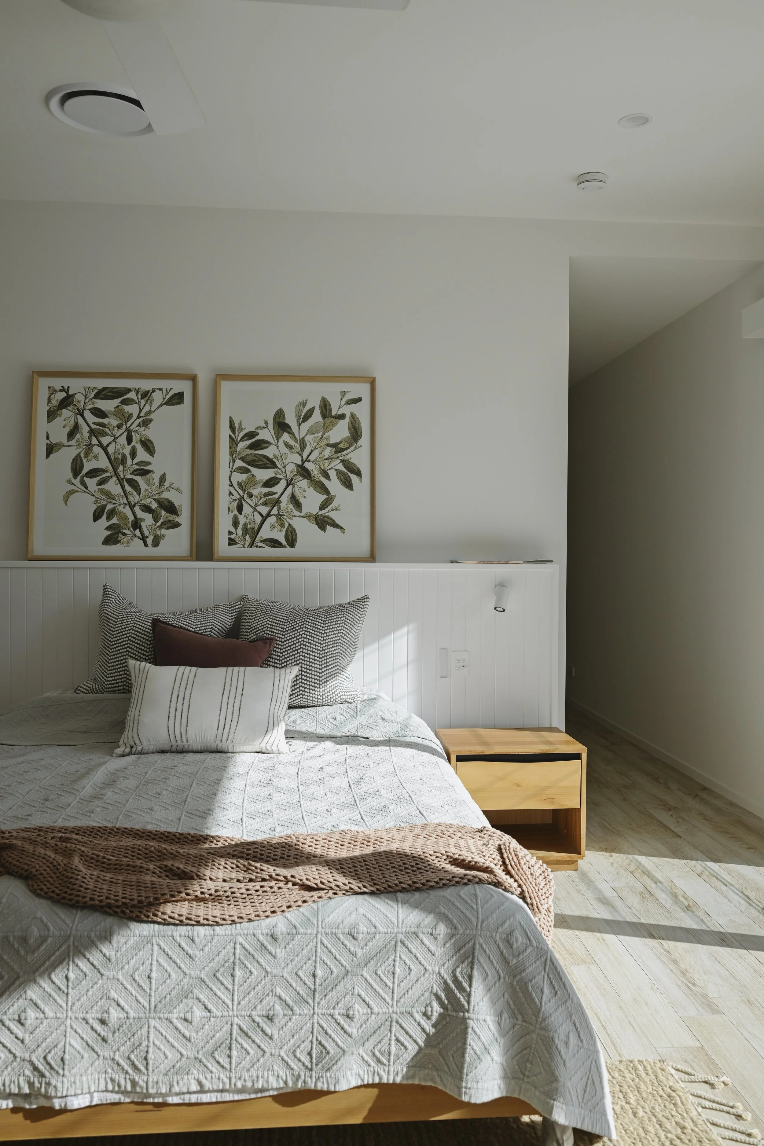 A cozy bedroom with a bed covered in a white quilt, decorated with various striped and patterned pillows. Above the bed, there are two framed botanical prints featuring green leaves. To the right of the bed, a small light wood nightstand with an open