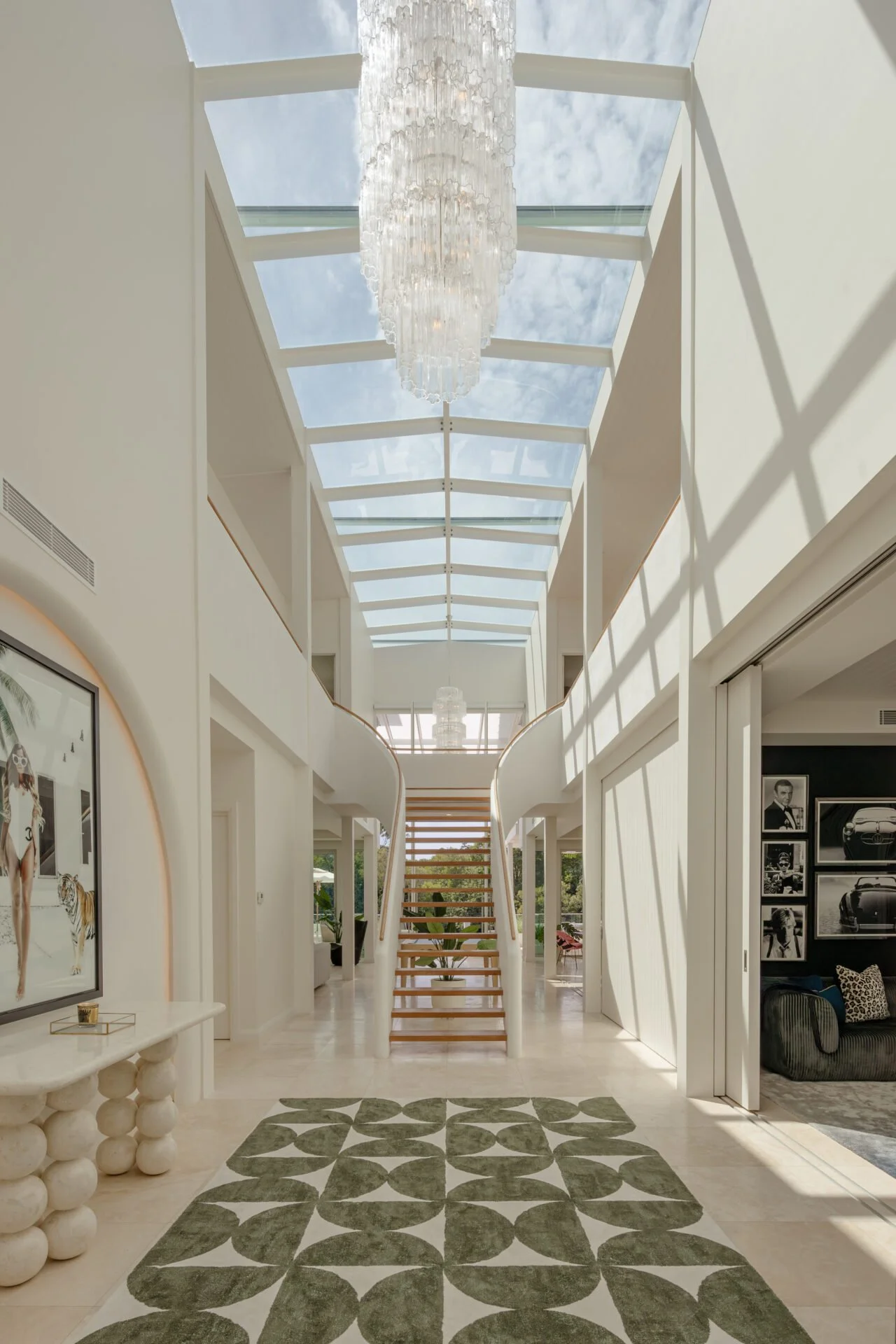 Bright modern hallway with a glass ceiling, a hanging chandelier, a staircase with open risers, and artwork on the walls.