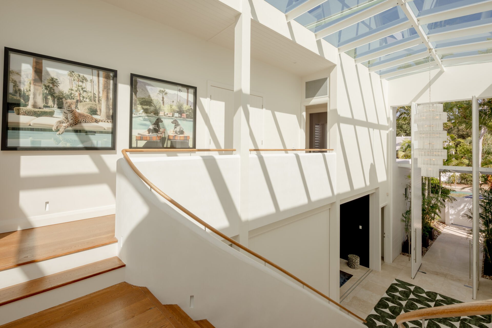 Bright indoor space with high glass ceiling, two framed pictures of animals on the wall, a staircase with wooden steps and a wooden handrail, and large open windows revealing lush trees outside.