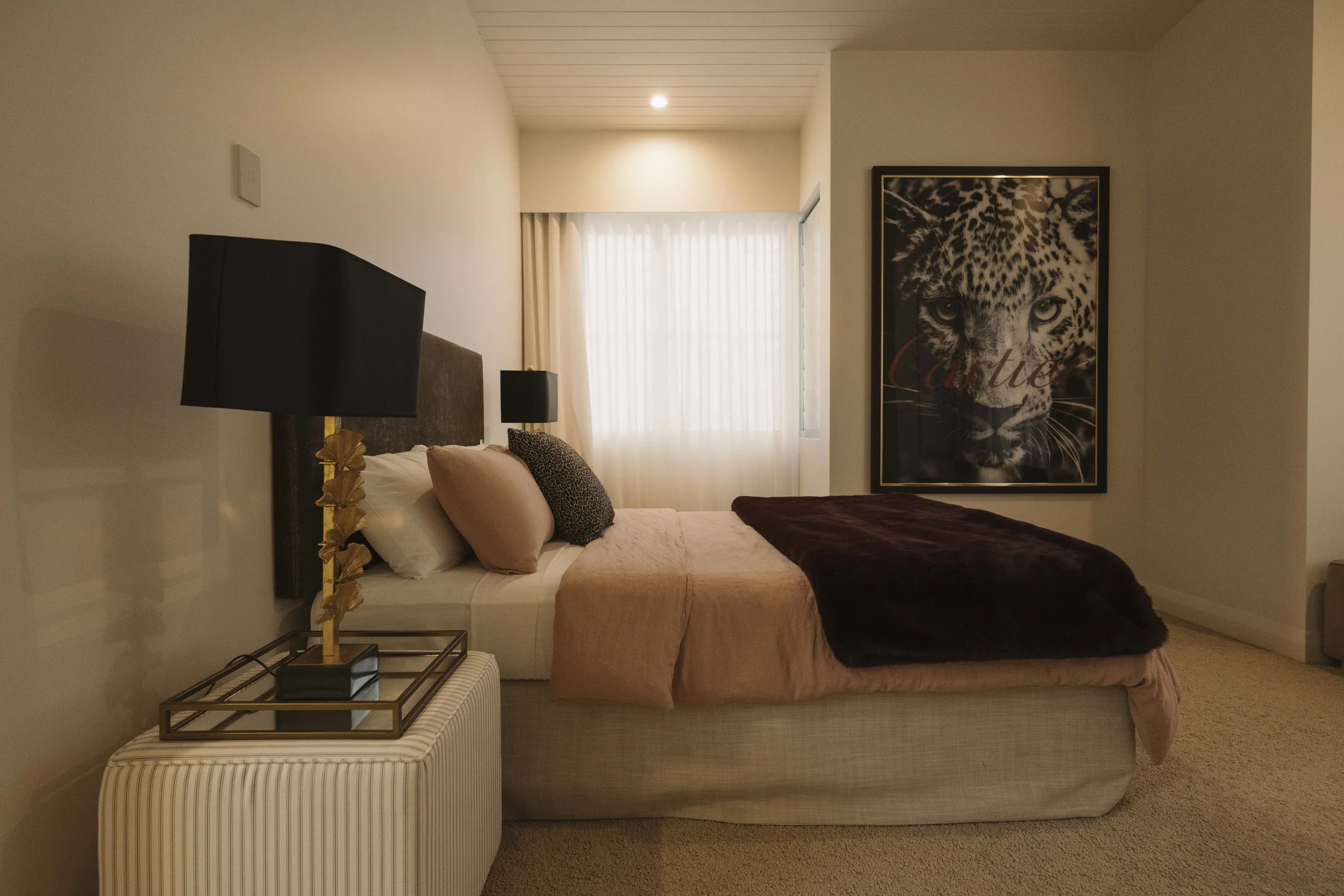 A bedroom with a bed featuring a brown blanket, pink and leopard print pillows, and a dark headboard. Two black lamps with golden bases sit on nightstands on either side of the bed. To the right, a large framed black-and-white picture of a leopard's 