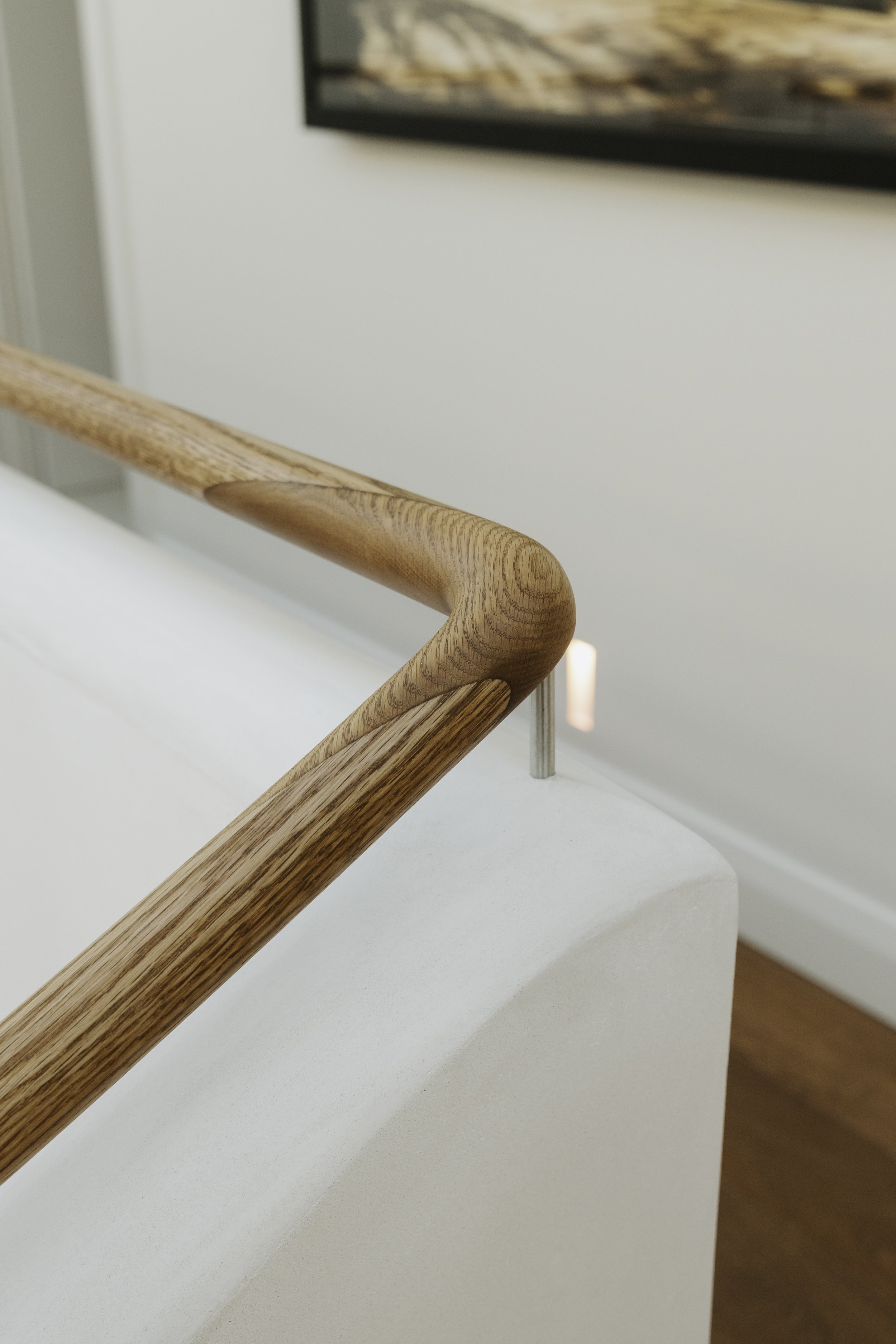 Close-up of a wooden handrail in a modern interior, with a white wall and part of a mounted picture frame in the background.