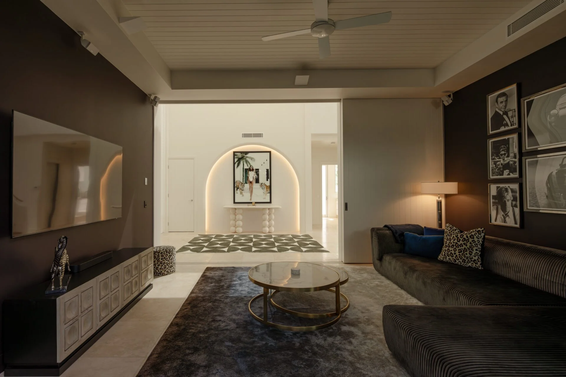 Modern living room with dark and light walls, featuring a black and white striped sofa, a leopard print pillow, a magazine on the side table, and a gallery wall of black and white photos. The room has a ceiling fan, floor lamp, and a rug, with a door