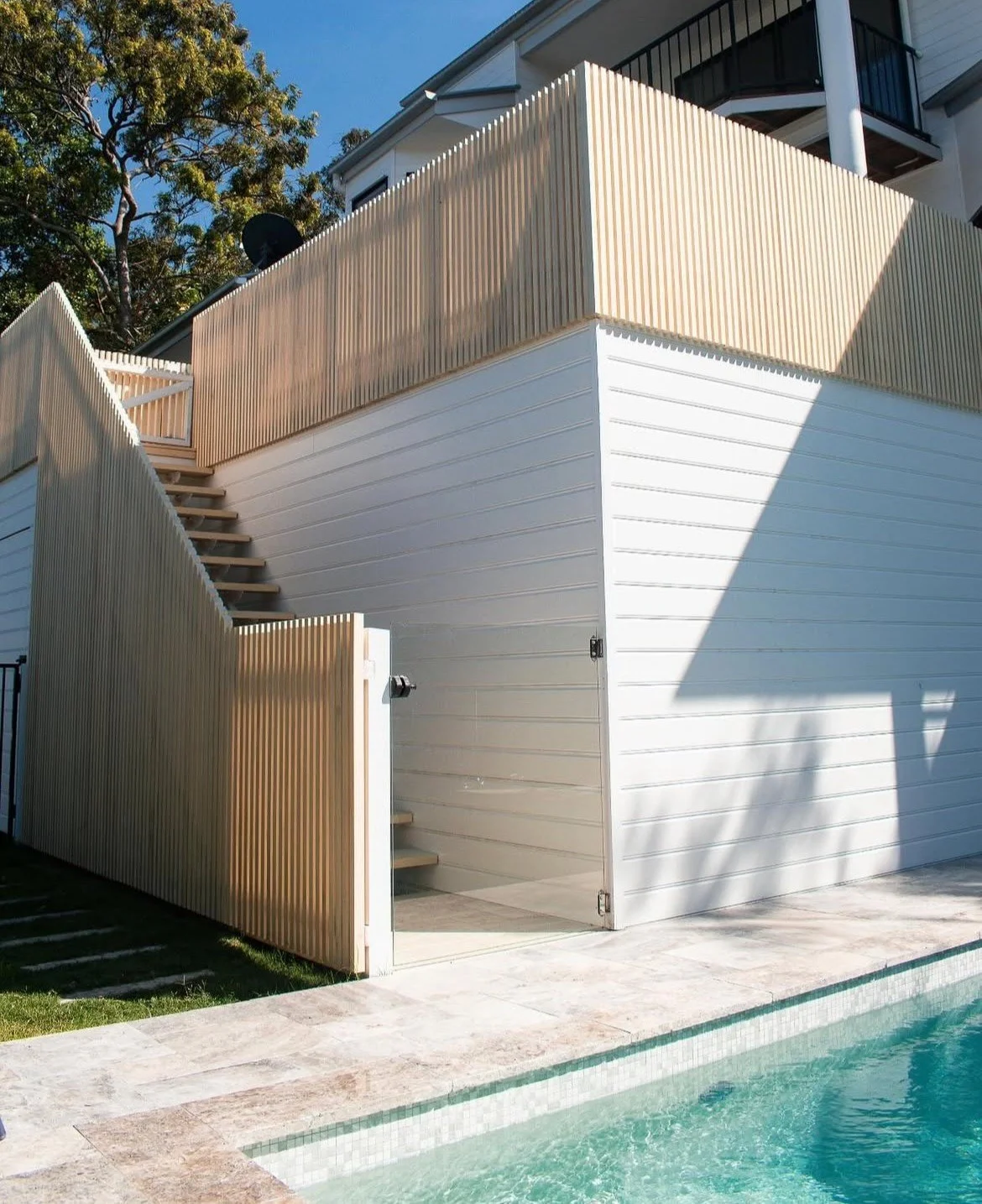 Exterior view of a modern house with a white wall, wooden fencing, open staircase, and a swimming pool in the foreground. Trees are visible in the background.