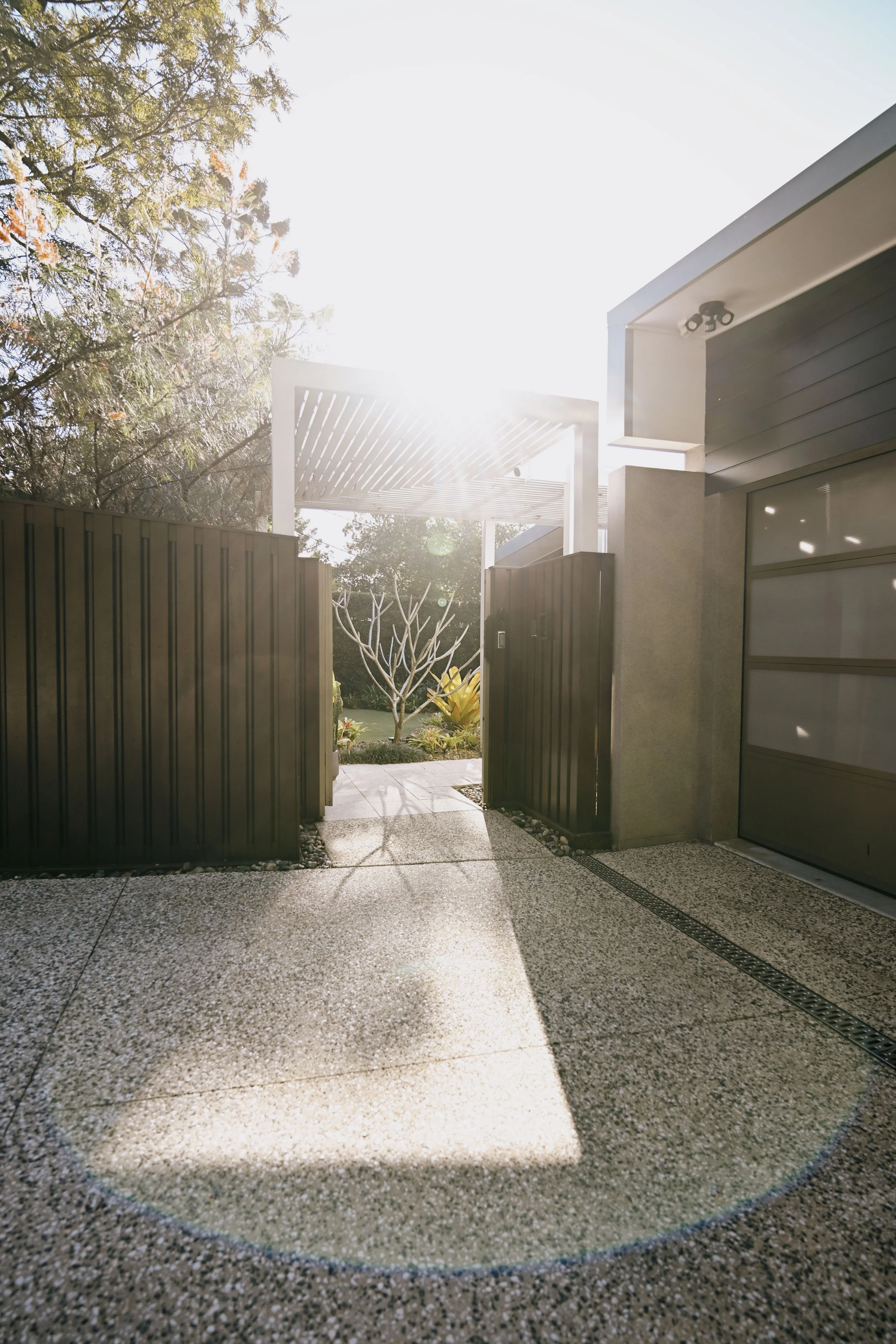 An outdoor view with an open gate leading to a garden area, sunlight shining brightly in the sky, trees and plants visible, and a modern house exterior with a garage door.