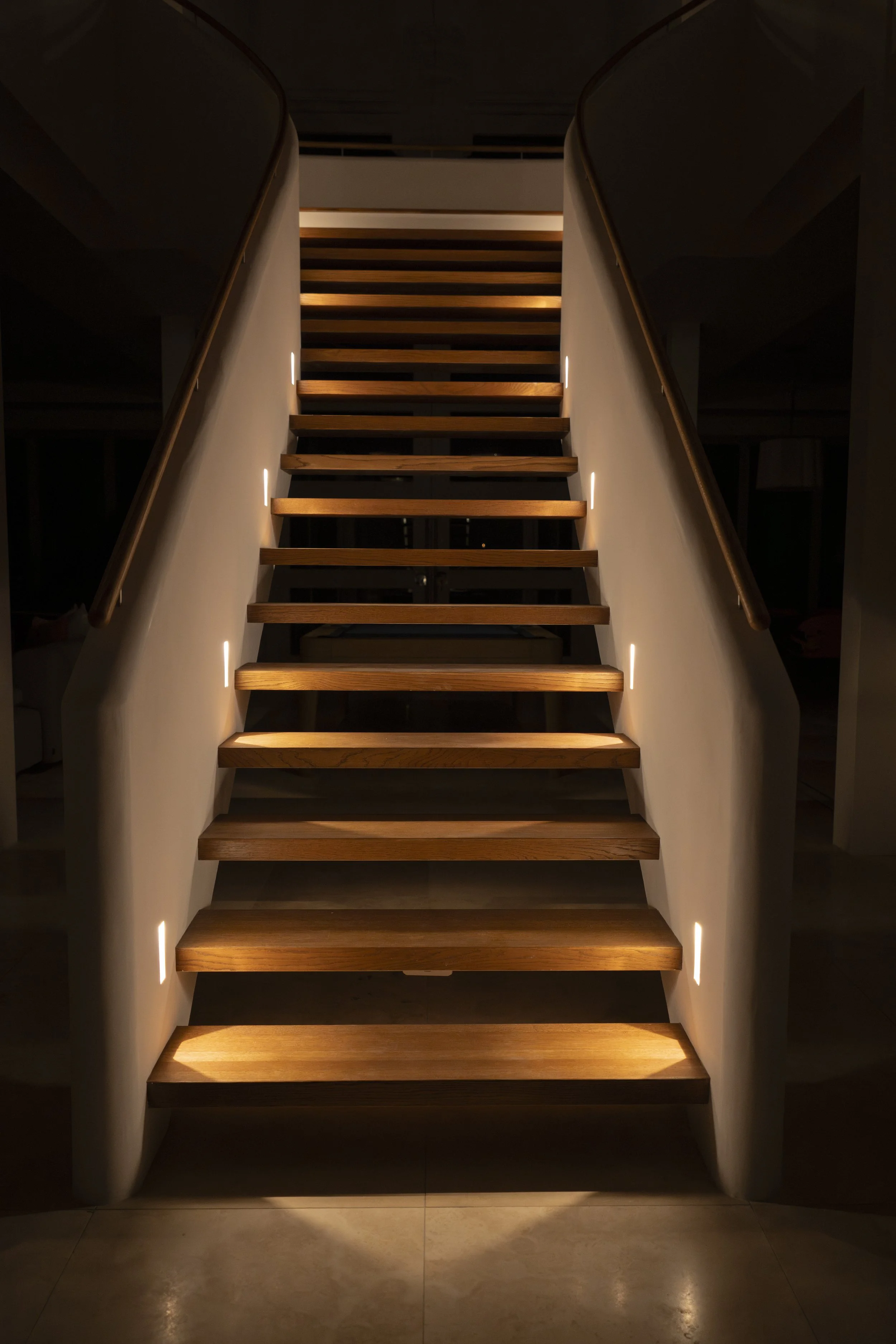 A staircase with wooden steps and white sides, illuminated by small lights along the walls, leading upwards.