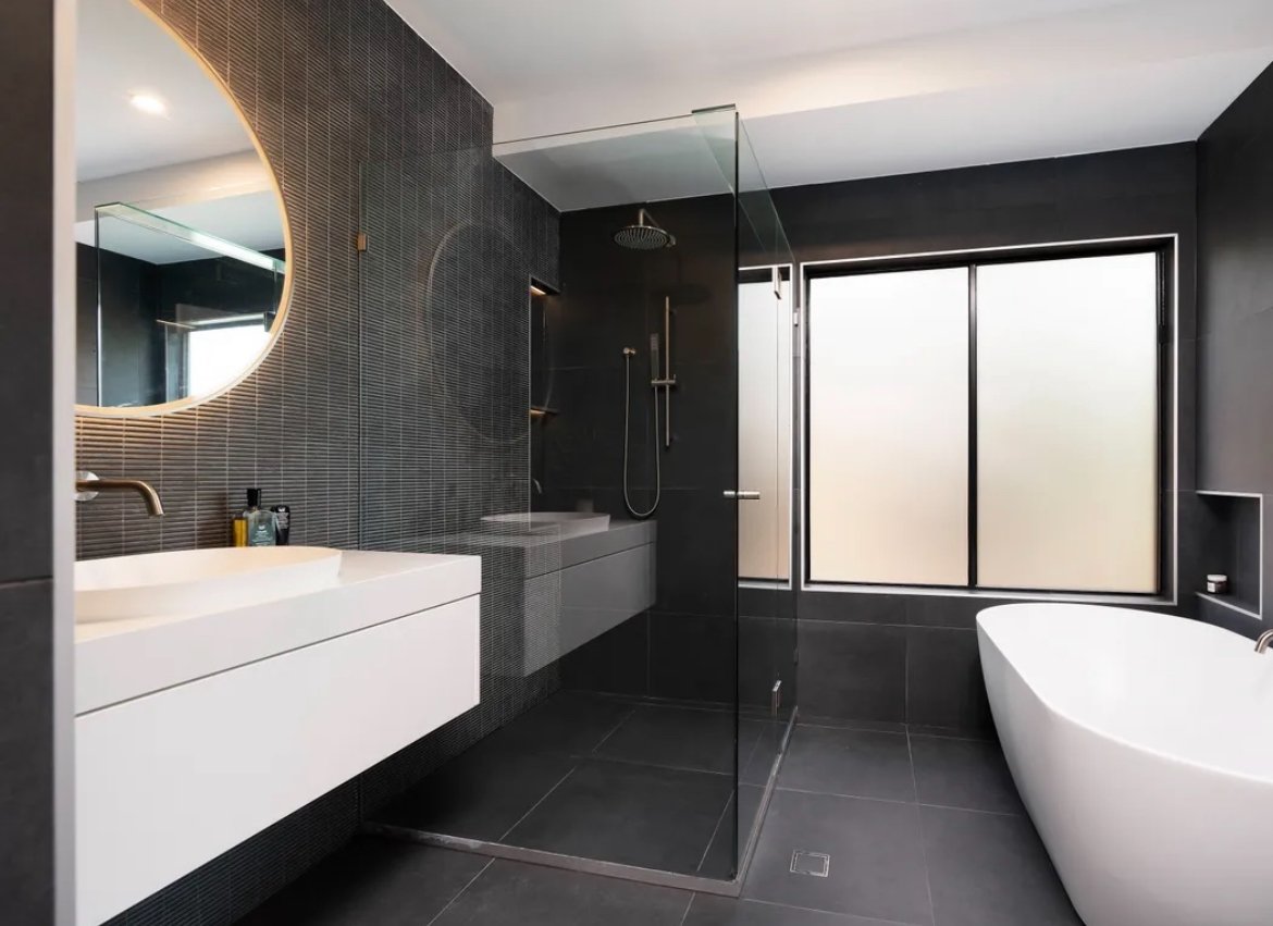 Modern bathroom with black walls and floor, a large window, a white bathtub, a floating vanity with a sink, a round mirror, and a glass shower enclosure.