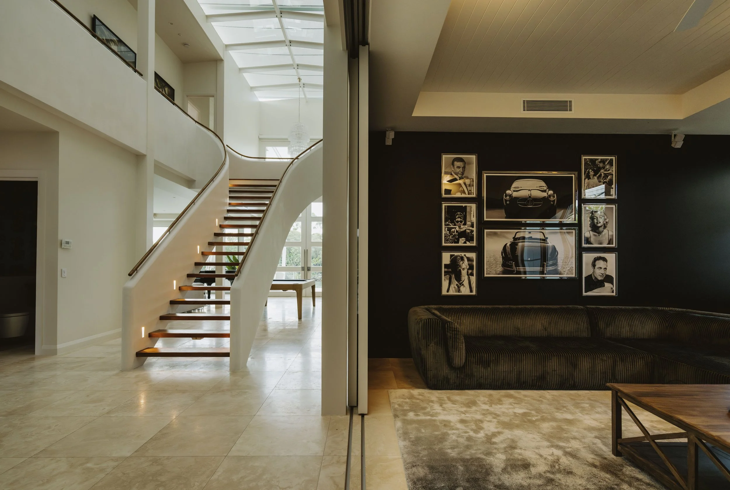 Interior view of a modern house with a grand staircase, black accent wall with framed vintage photographs and artwork, cream-colored marble floor, and a large skylight ceiling with a chandelier.