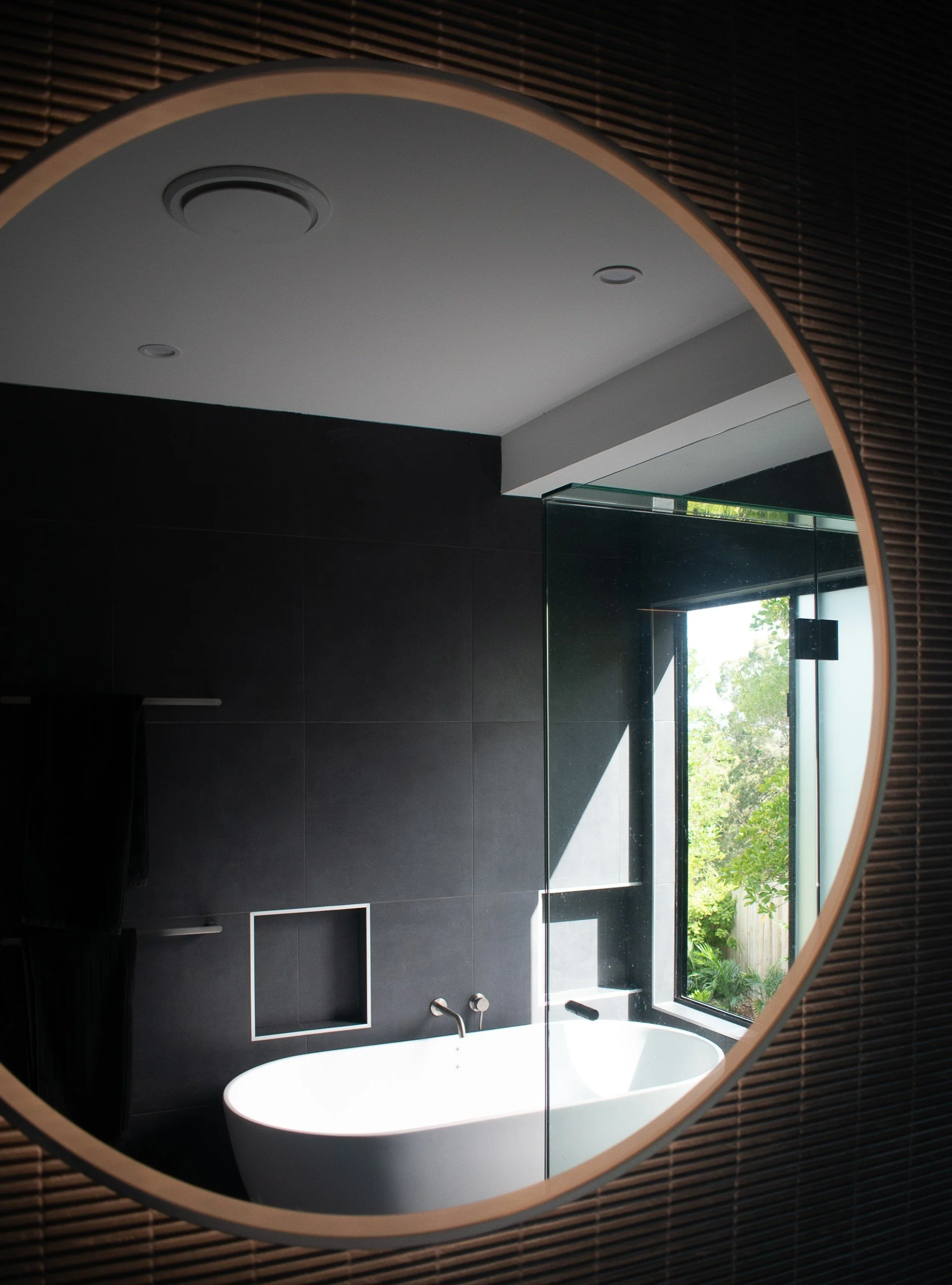 Modern bathroom with black tiled walls, a built-in rectangular shower niche, a white bathtub, and a large window with outdoor greenery visible.