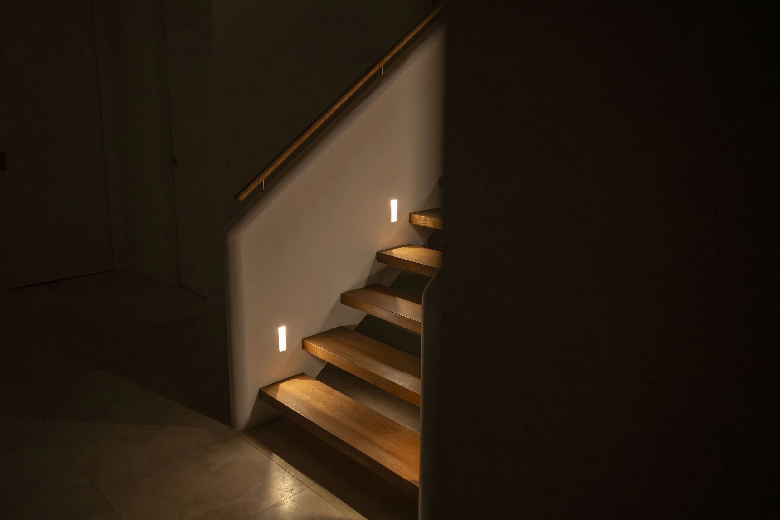 A dimly lit staircase with wooden steps and a black wall on one side, illuminated by small rectangular wall lights.