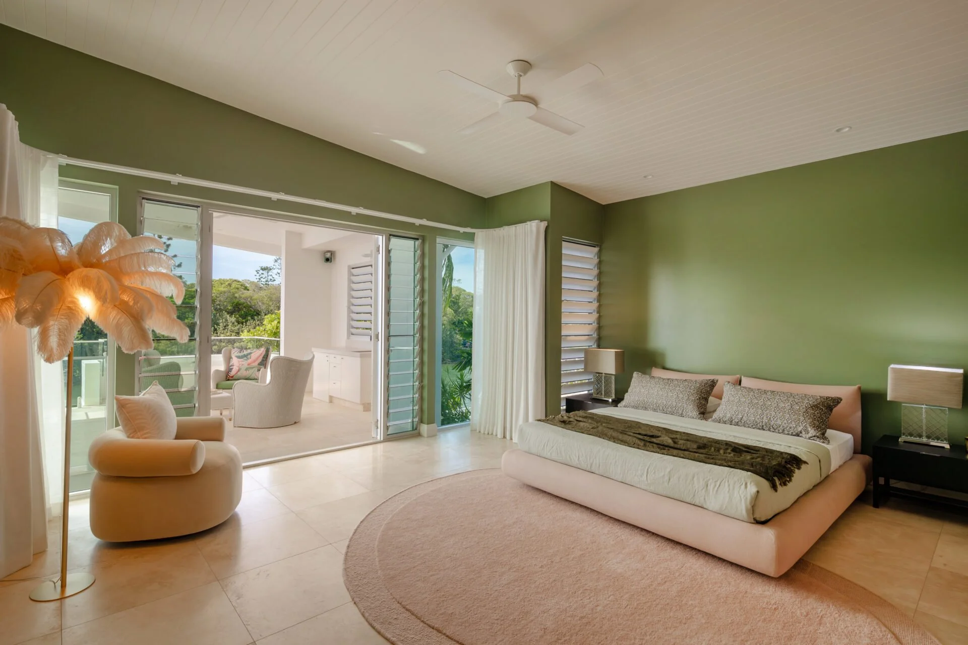 Living room with green accent wall, beige sofa, white armchair, lamp with feather shade, round beige rug, large glass sliding door leading to balcony, view of trees outside.