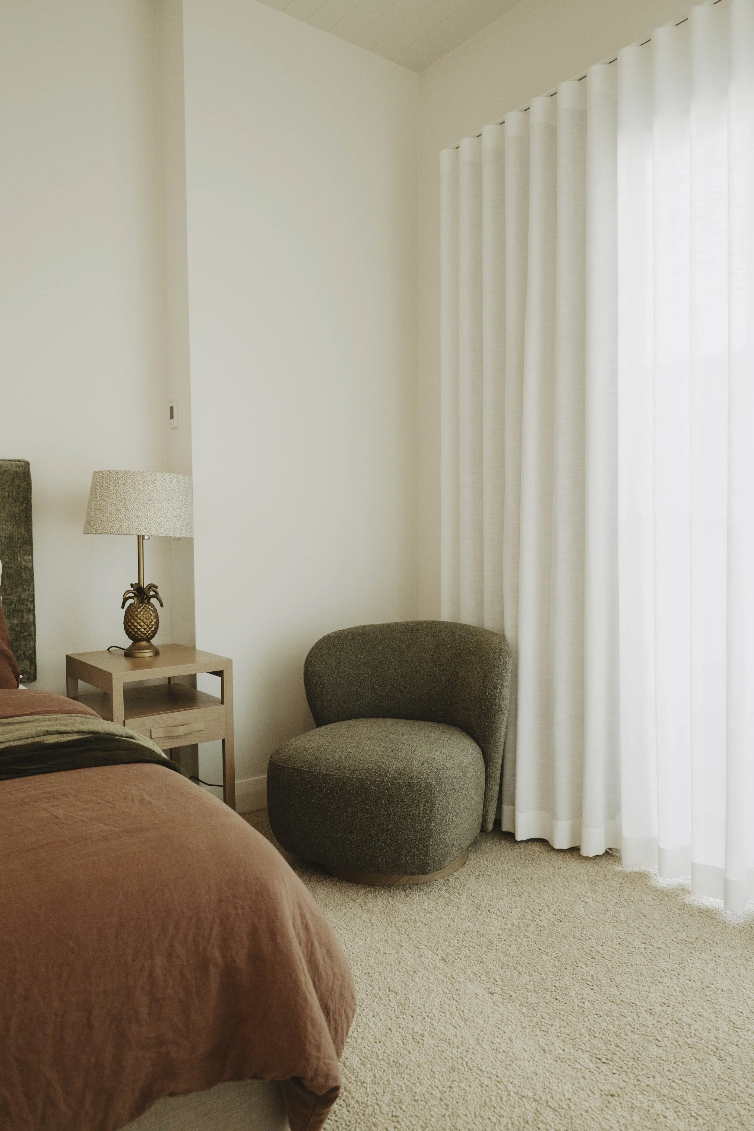 A bedroom corner with a bed, a nightstand, a lamp with a pineapple-shaped base, a gray upholstered chair, and white curtains covering a window.