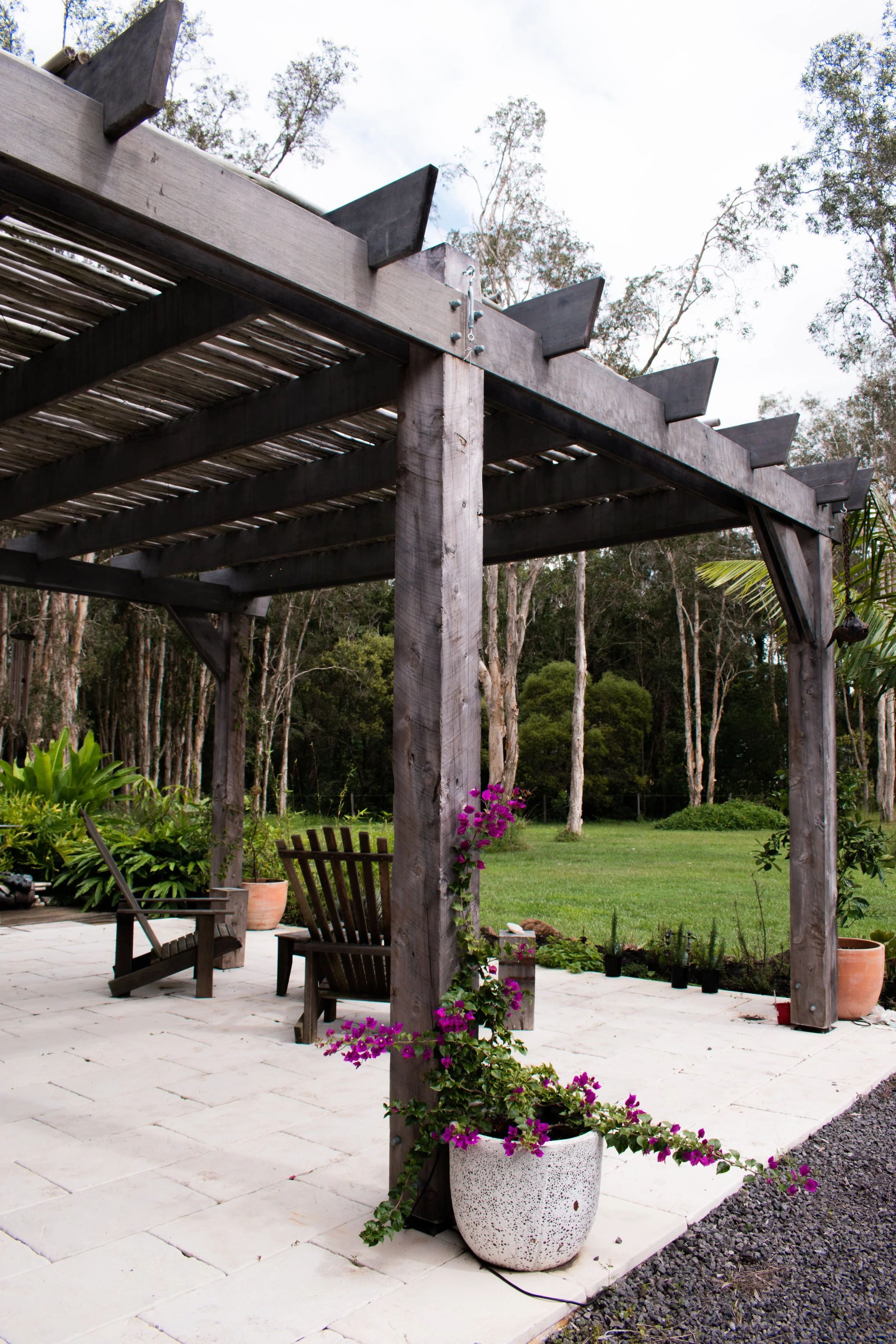 A backyard patio with a pergola, overlooking a grassy yard with trees. There are potted plants and garden furniture, with pink flowering plants climbing on a wooden post in the pergola.