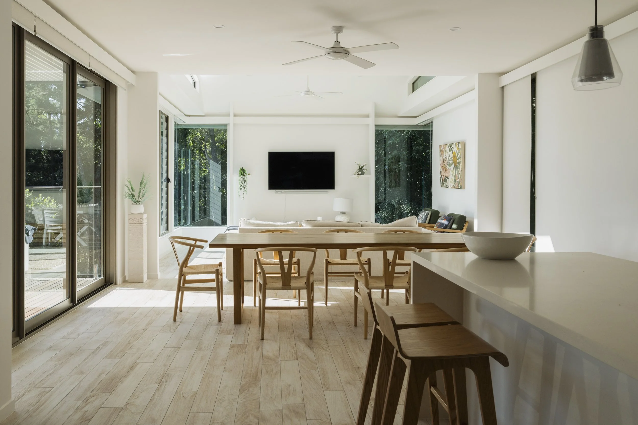 Bright open-concept living and dining area with large glass sliding doors to the balcony, light wood flooring, white walls, and minimalist decor including a wall-mounted flat screen TV, a ceiling fan, and a few potted plants.