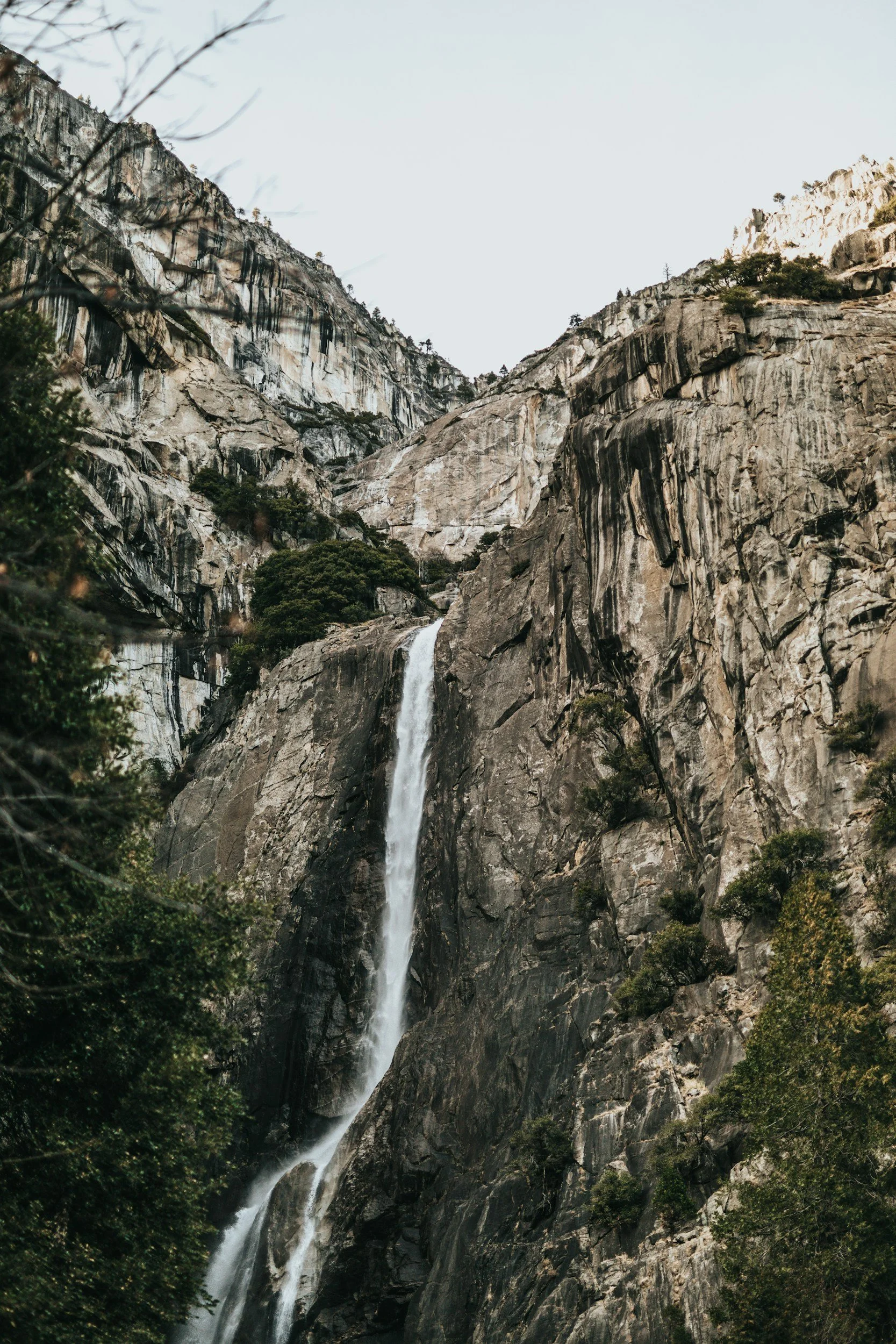 A tall mountain waterfall flowing down a rugged cliff through sparse trees.