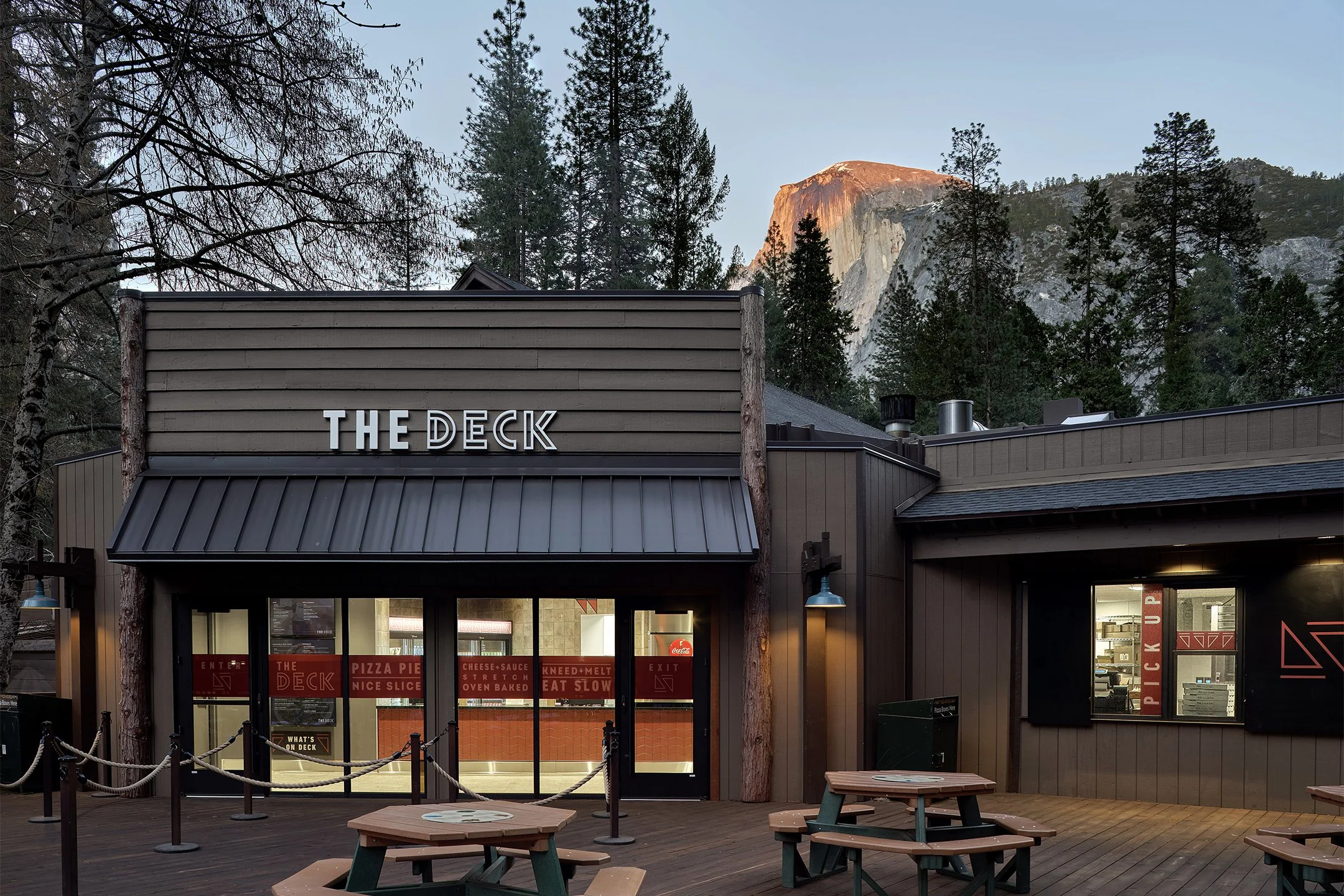 Exterior of a restaurant called The Deck with picnic tables on a wooden deck, surrounded by trees and mountains in the background.