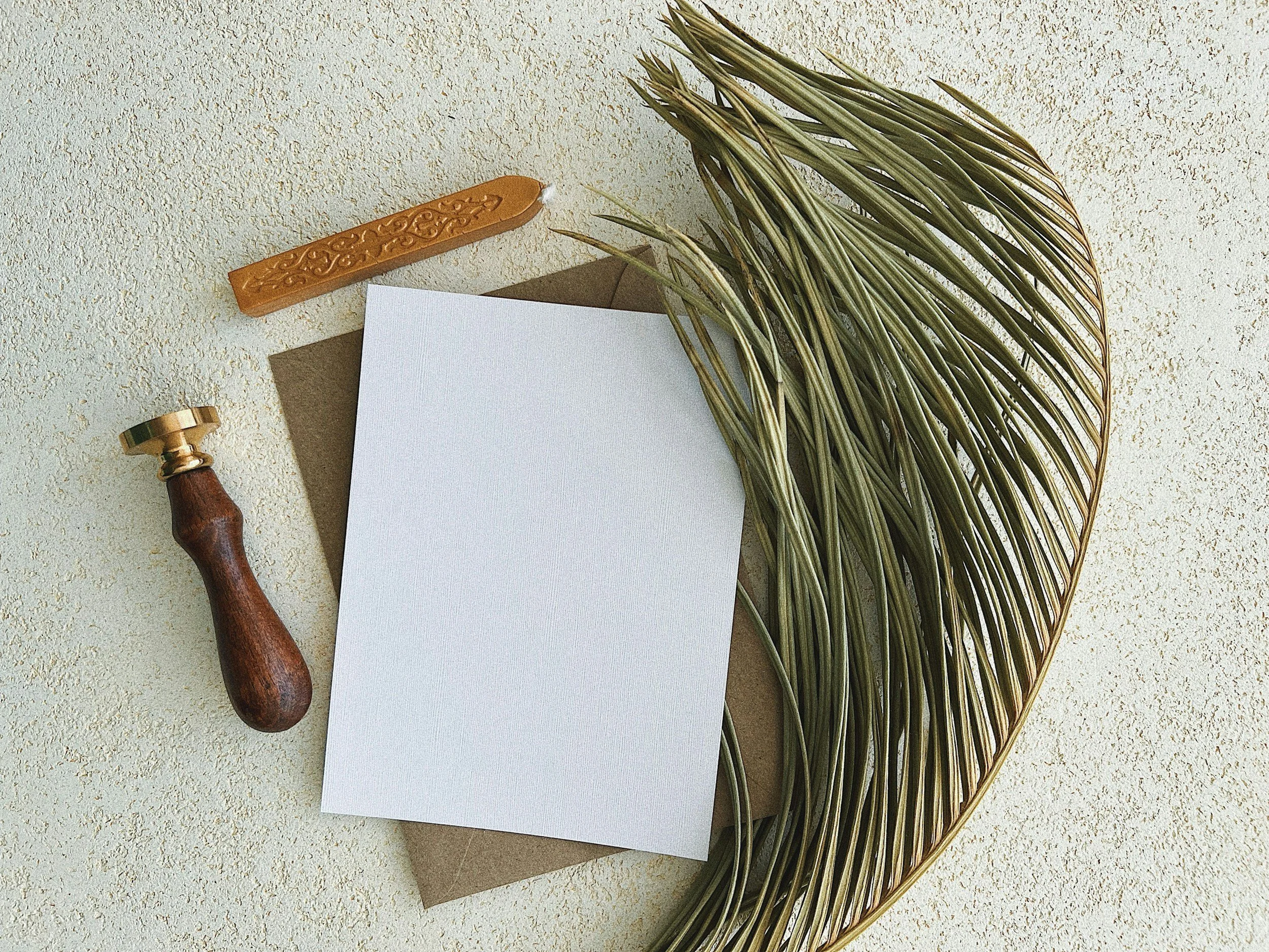 Flat lay of a white blank paper, a brown envelope, a wooden-handled wax seal stamp, a brown wax stick, and a large decorative gold wire leaf on a light textured surface.