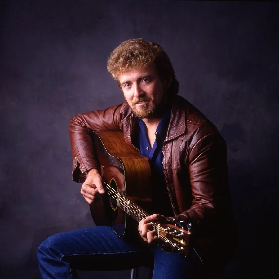 Keith Whitley with curly hair and a beard wearing a brown leather jacket, sitting and playing an acoustic guitar, against a dark background.