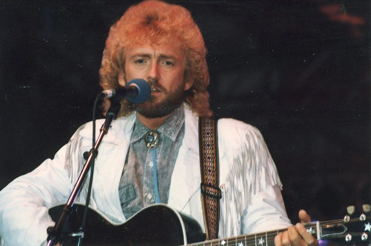 A man with curly red hair and a beard singing into a microphone while playing an acoustic guitar.