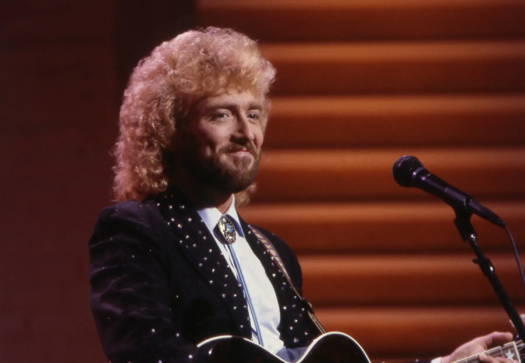 Keith Whitley with curly blond hair and a beard, wearing a black jacket with sparkles, a white shirt, and a bolo tie, stands in front of a microphone holding a guitar.
