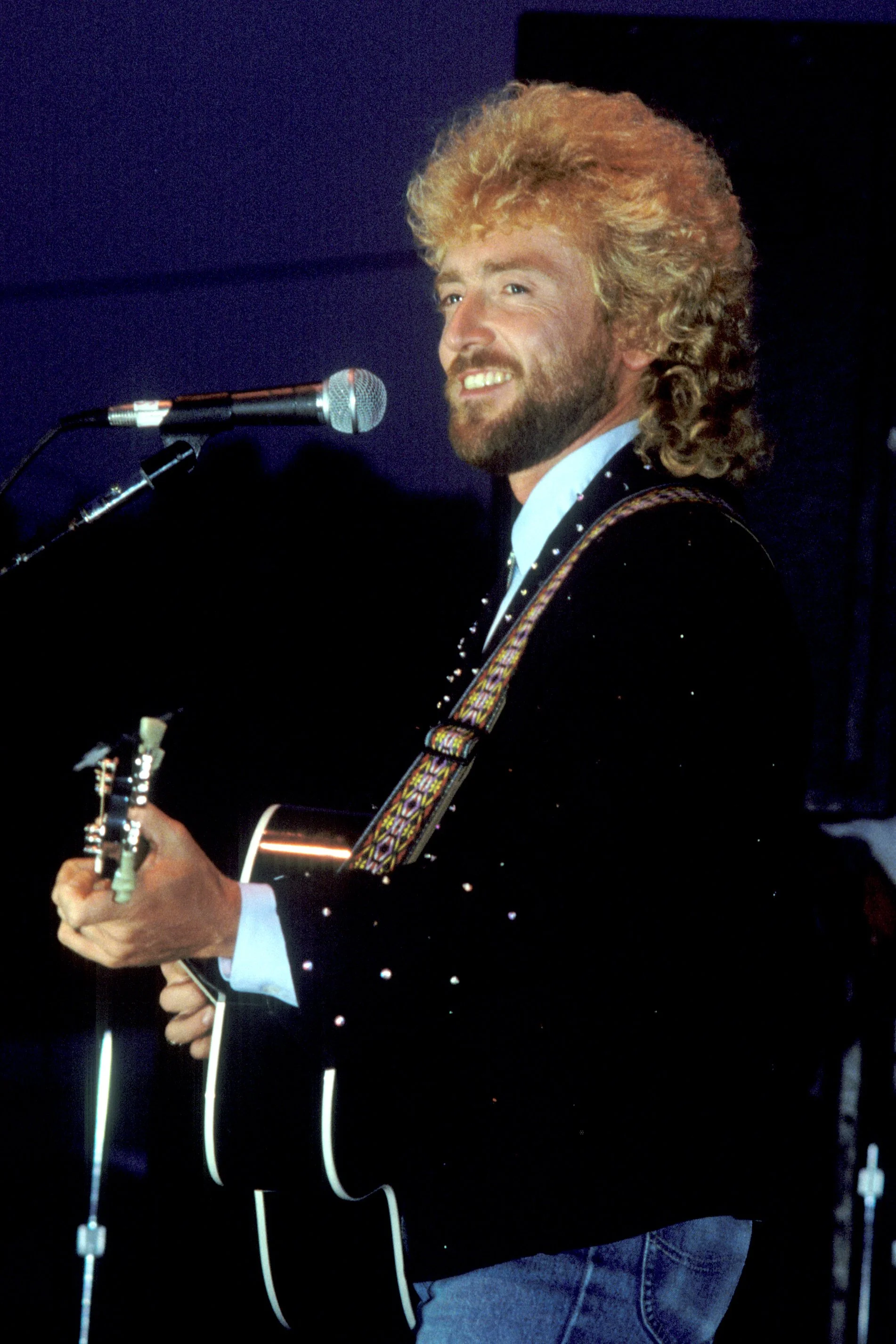 Keith Whitley with curly blond hair and a beard playing an acoustic guitar and singing into a microphone.