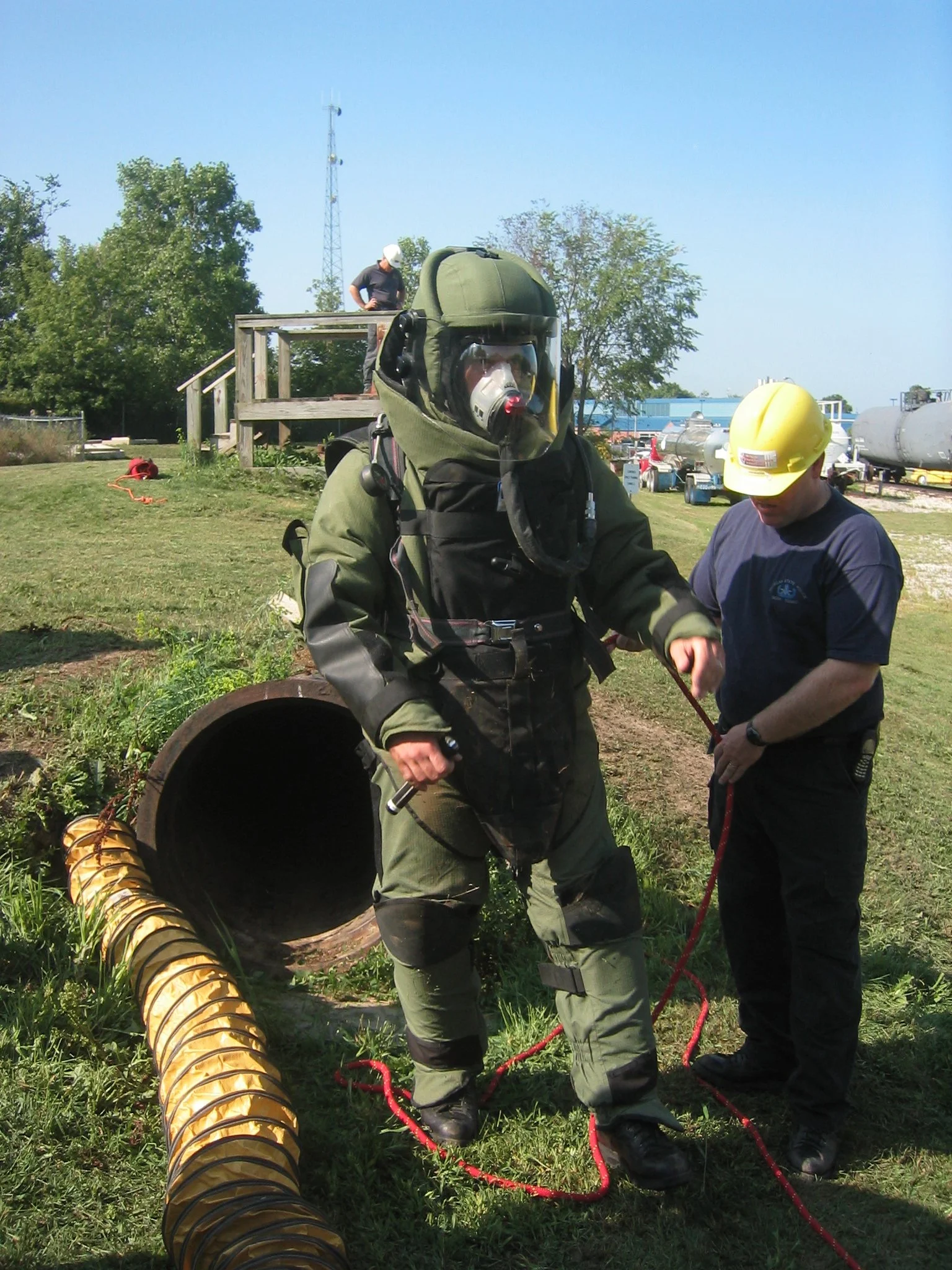 A firefighter in protective gear with a face mask and oxygen tanks being assisted by a man in a yellow helmet near a large pipe and yellow ventilation duct outdoors.