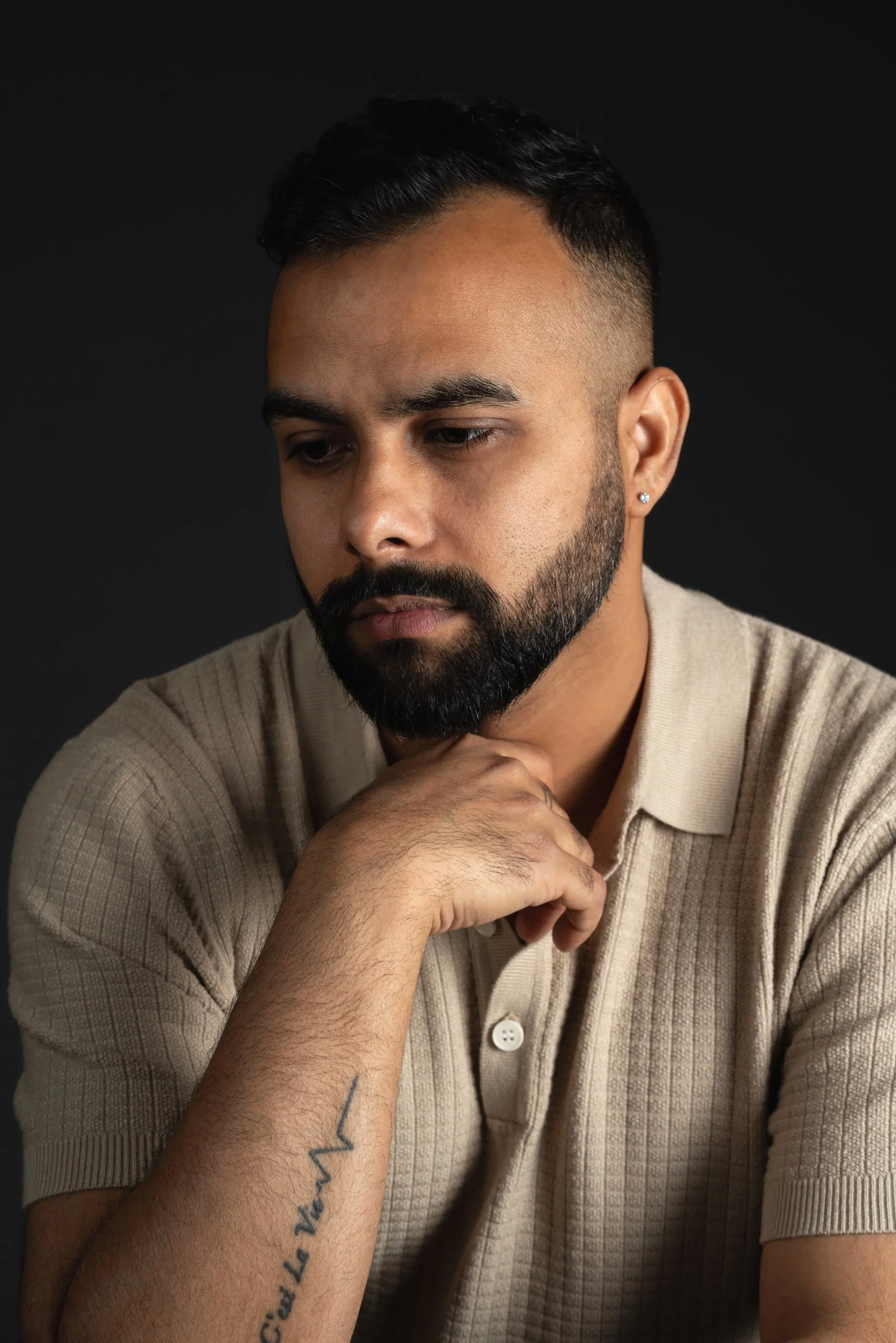A man with a beard and mustache, wearing a beige shirt, looking down thoughtfully against a black background.
