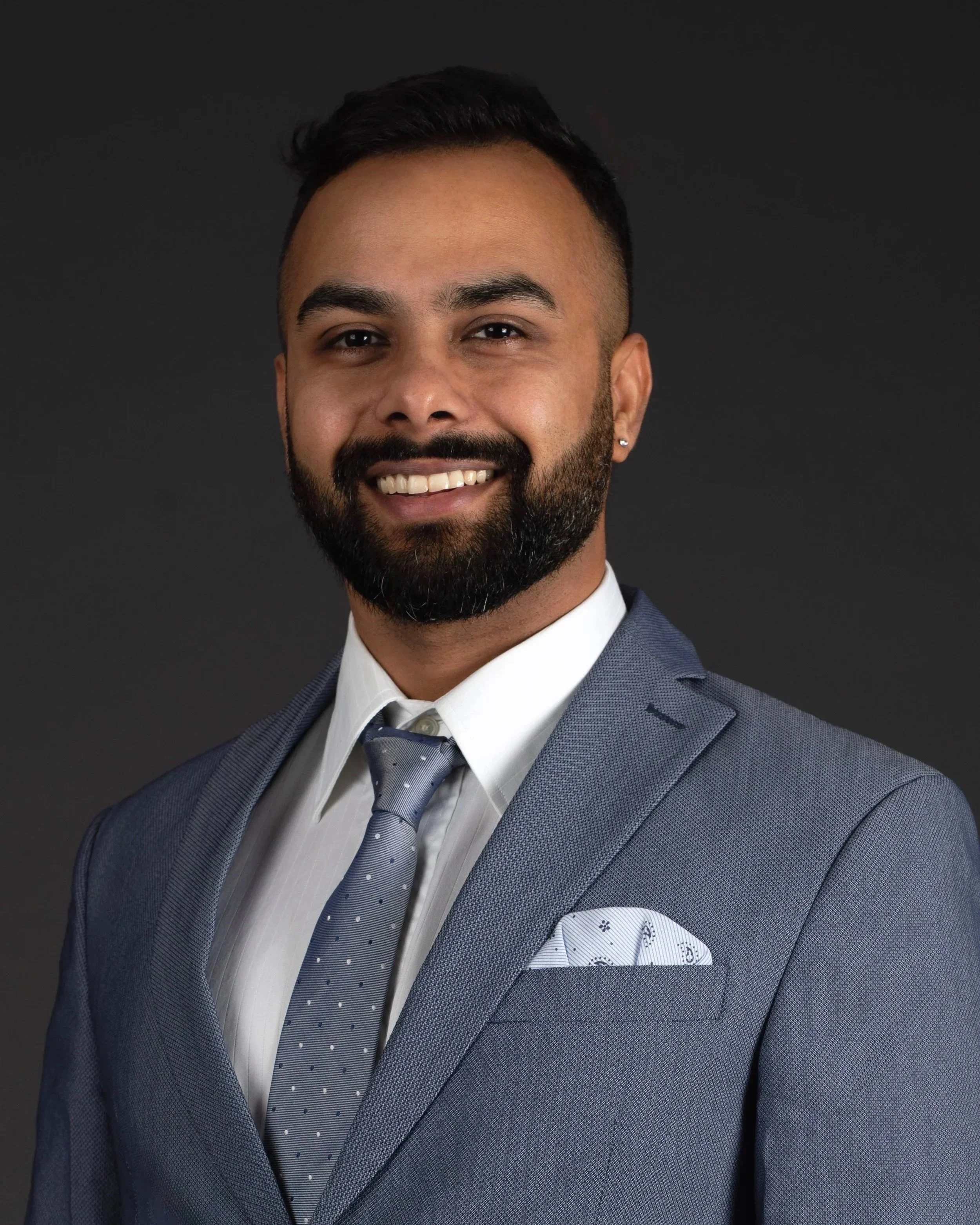 Professional headshot of a smiling man with a beard, wearing a gray suit and tie, with a white pocket square, against a dark background.