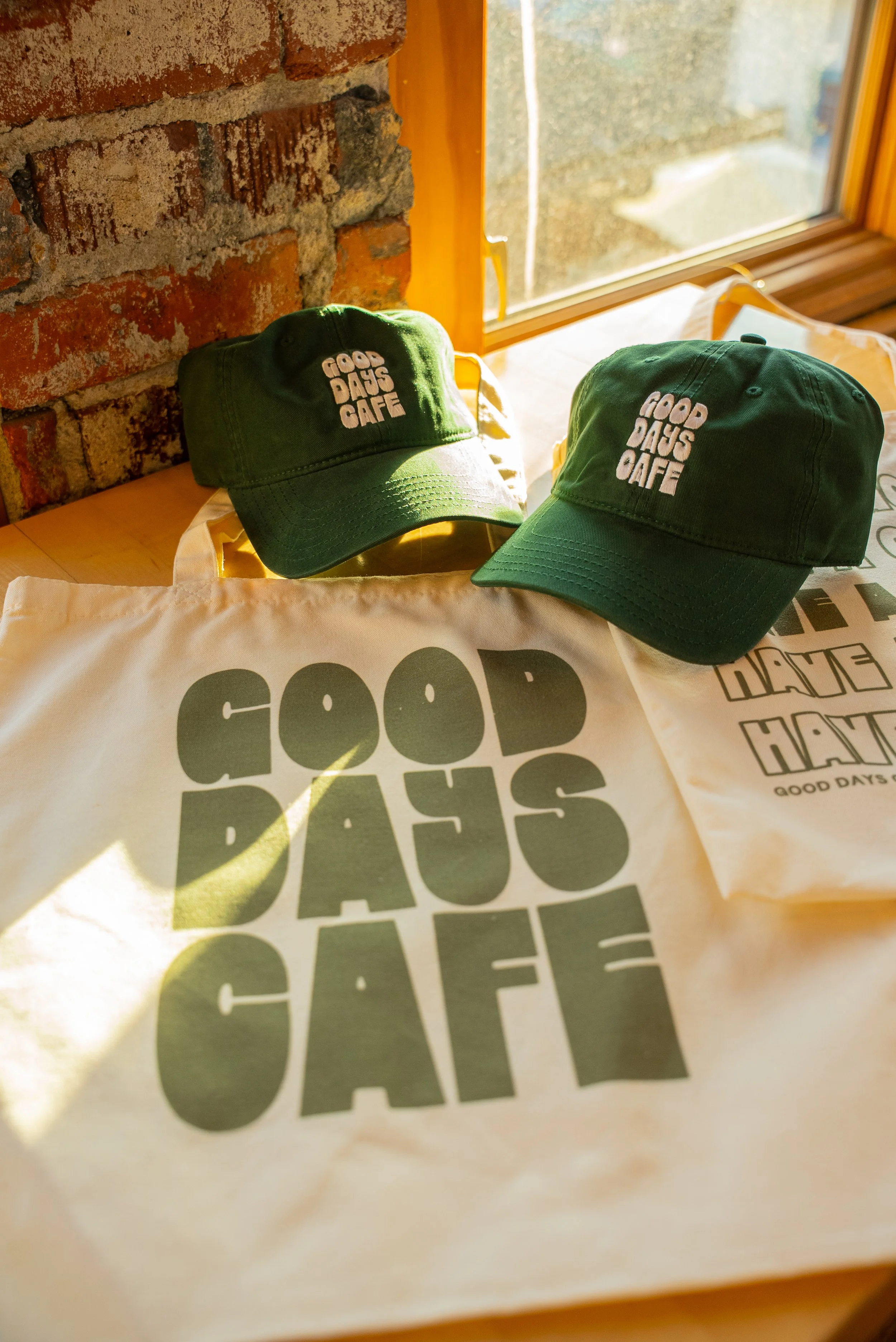 Two green caps and paper bags with 'Good Days Cafe' logo placed on a wooden table near a window with brick wall in the background.