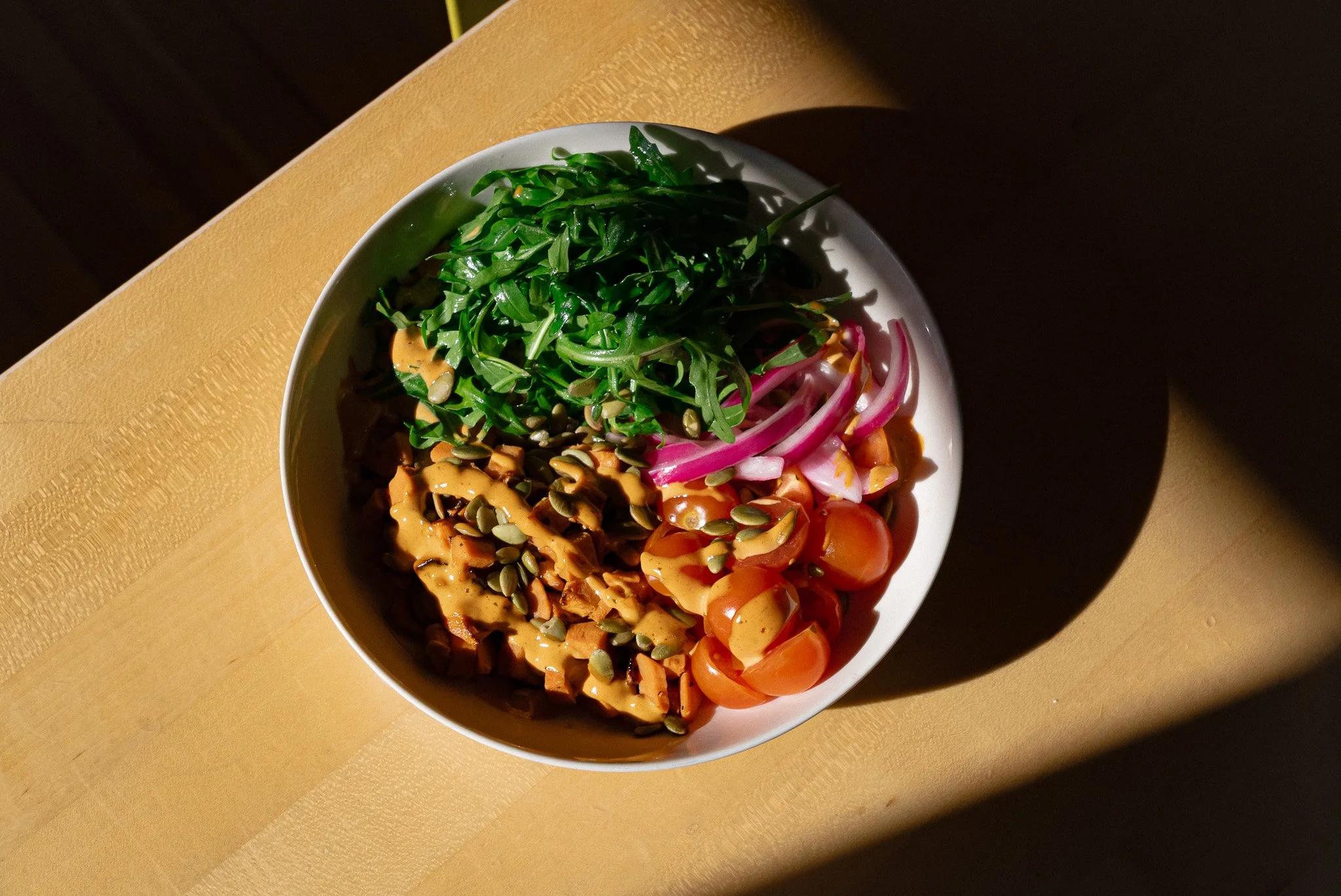 A bowl of salad containing cherry tomatoes, red onions, greens, and pumpkin seeds on a wooden table.