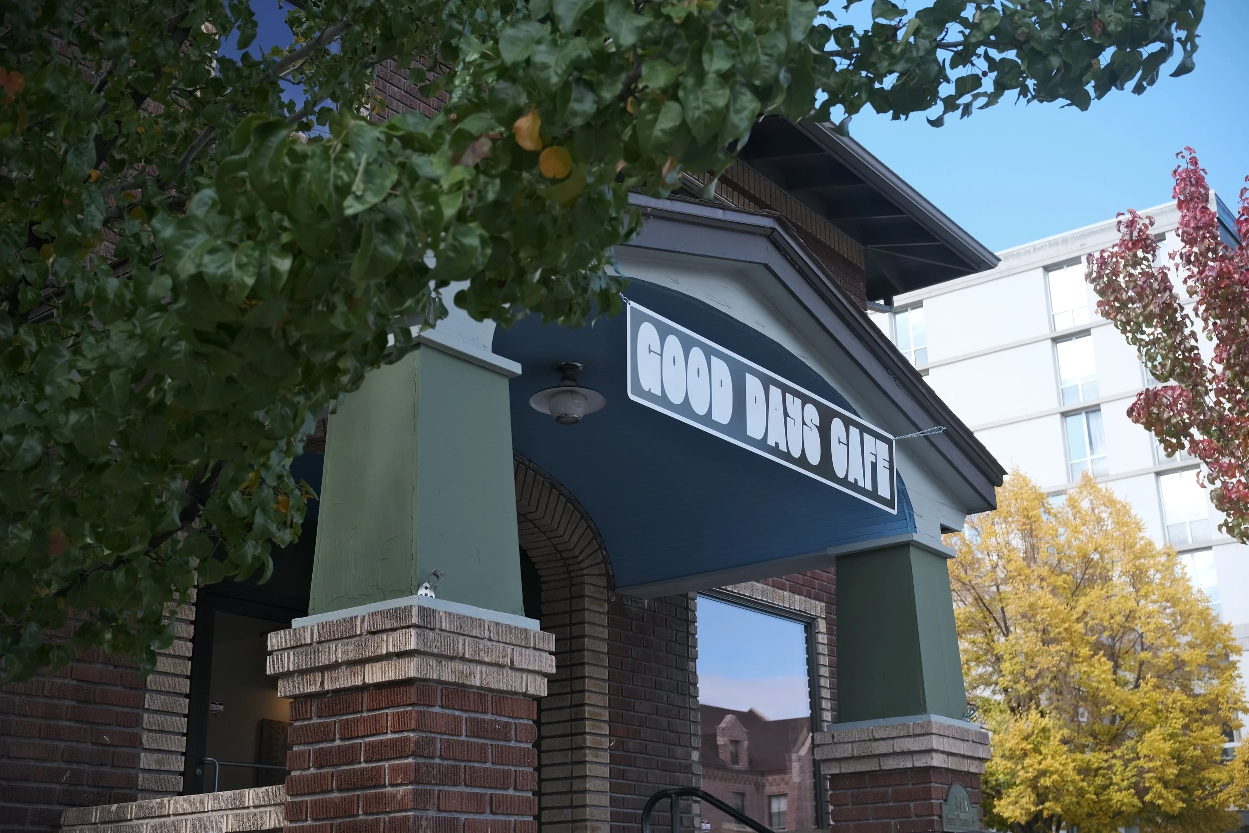 The exterior of a cafe named 'Good Days Cafe' with brick and painted wood architecture, surrounded by trees with green and yellow leaves, under a clear blue sky.