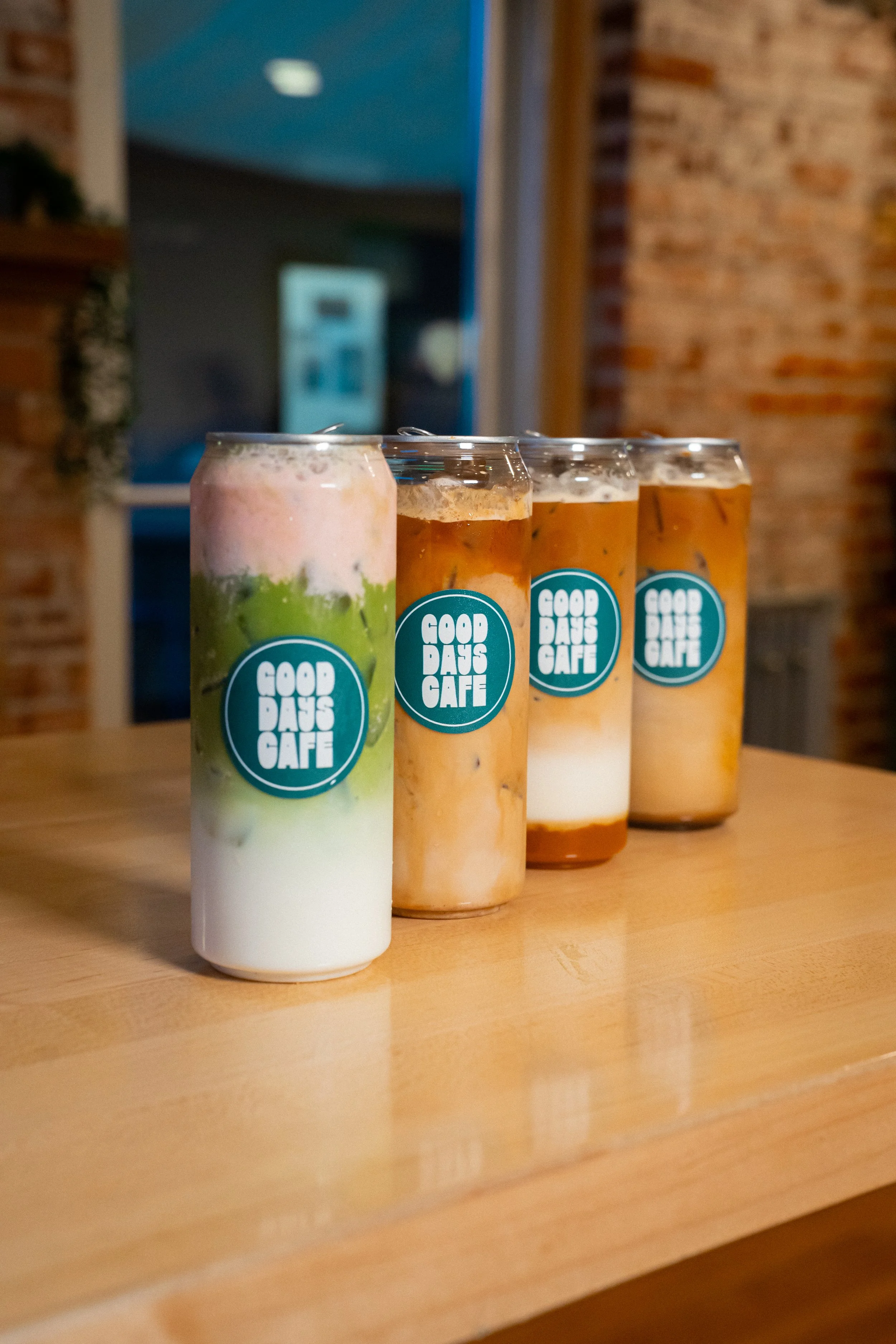 Four cans of beverages on a wooden table, with a brick wall in the background, labeled 'Good Days Cafe.'
