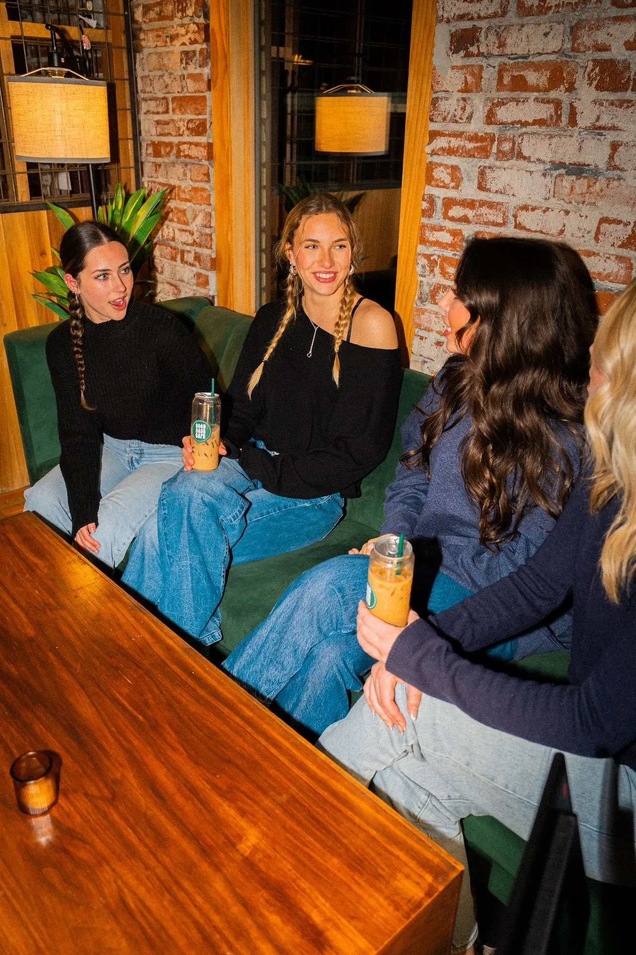 Four women sitting on a green couch at a wooden table in a cozy, brick-walled cafe, conversing and holding iced coffee drinks.