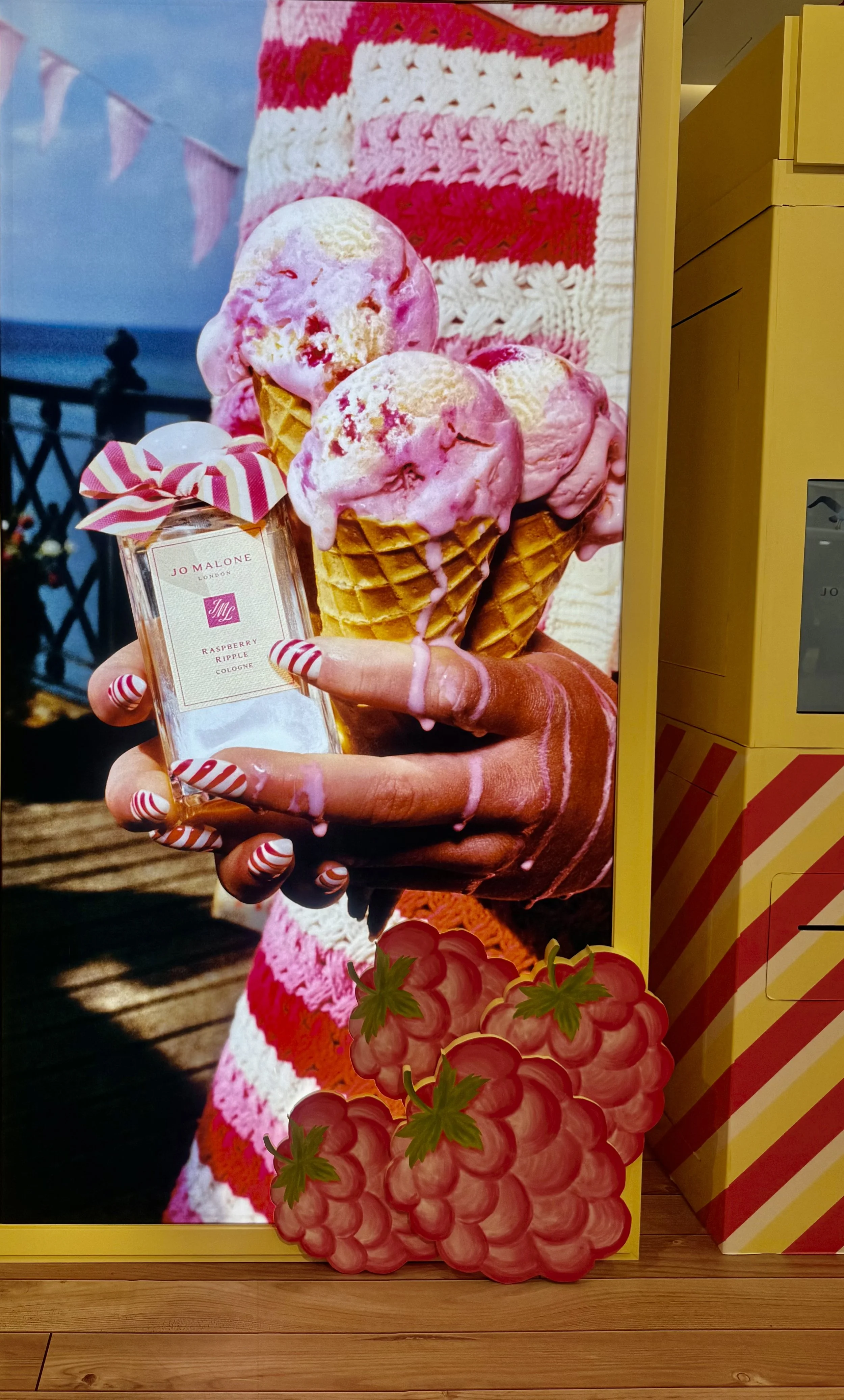 A poster showing three pink and white ice cream cones held by a hand with striped fingernails, in front of a background with pink and white striped fabric, a perfume bottle labeled 'Jo Malone Raspberry Ripple Cologne,' and illustrated raspberries at the bottom.