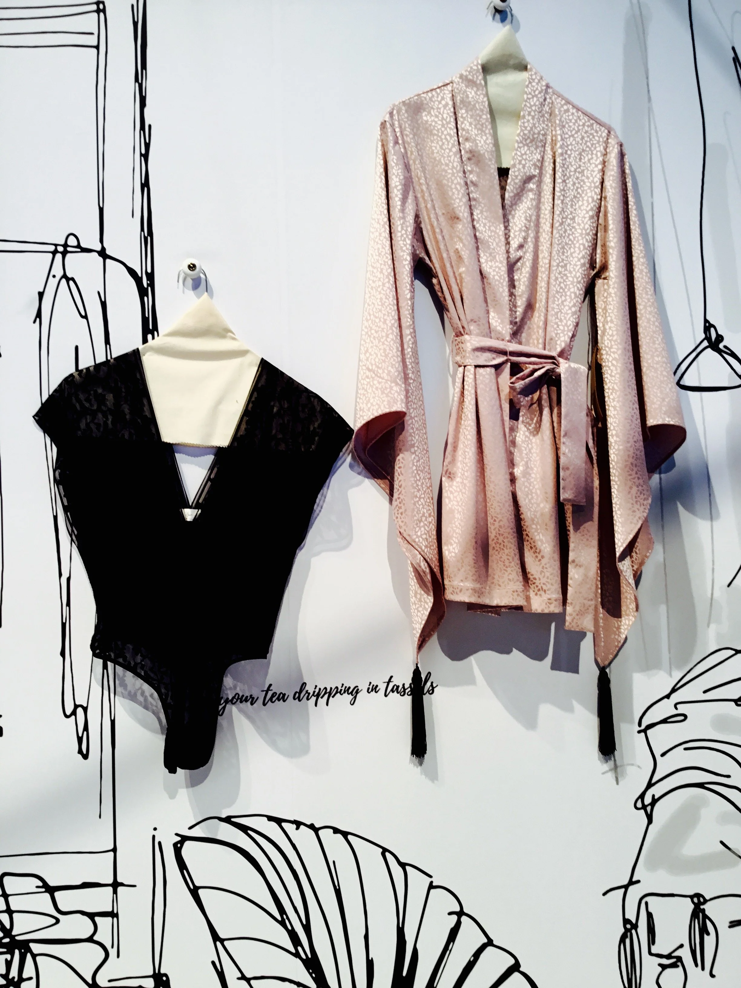 Two clothing items hanging on a wall: a pink satin robe with a belt and a black bodysuit with a beige collar.