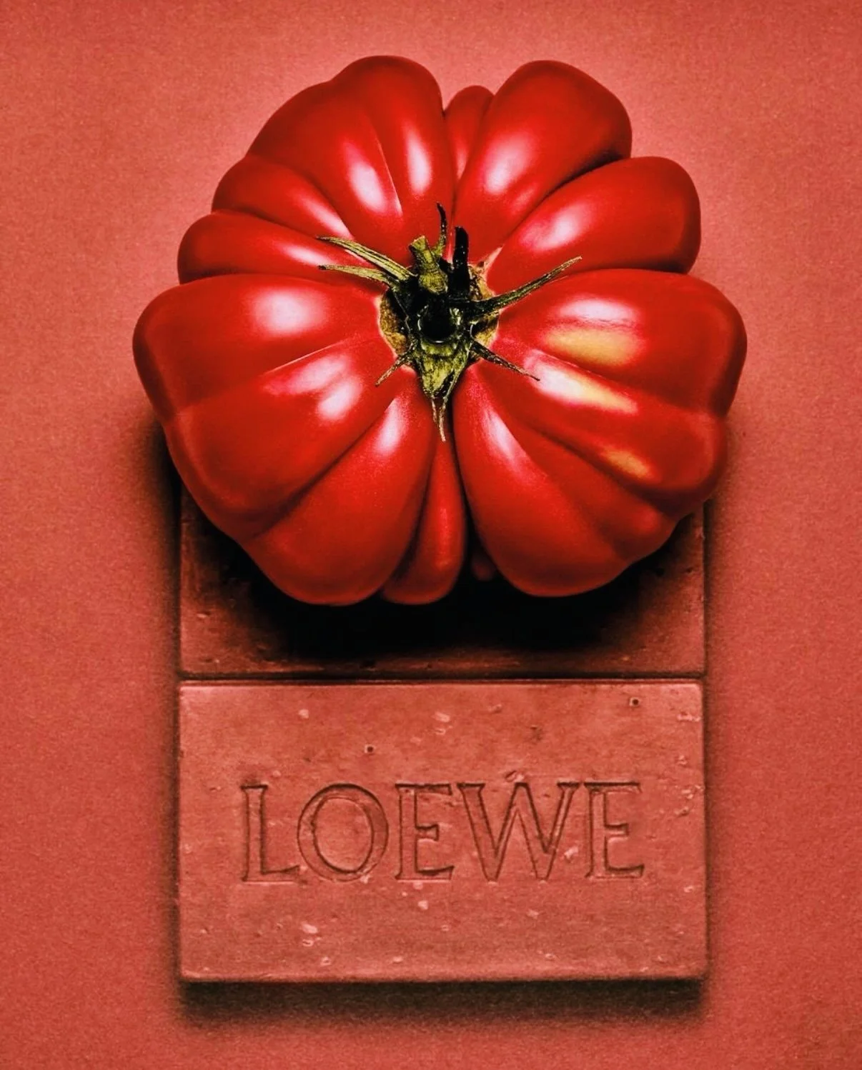 A large, plump, red heirloom tomato with ridges, placed on a square, rusty metal stand with the word "LOEWE" engraved, set against a red background.