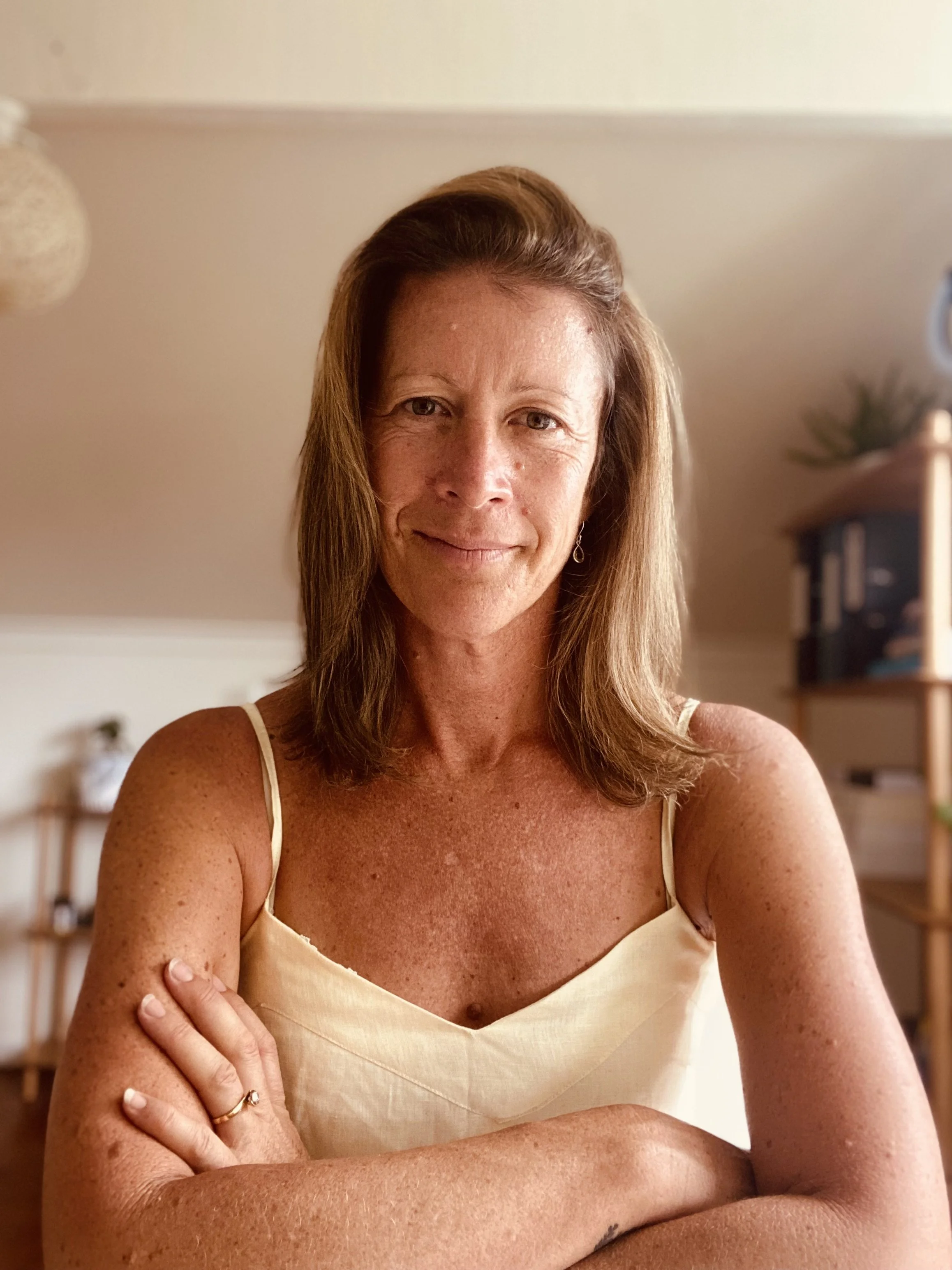 A woman with shoulder-length brown hair and freckles, wearing a yellow sleeveless top, smiling with arms crossed in a room with wooden shelves and indoor plants in the background.